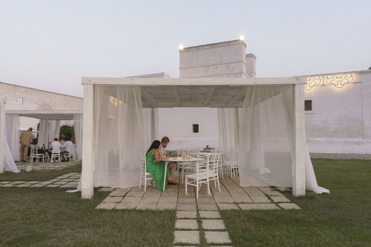White draped cabana with chairs and tables set up on grass at dusk for an outdoor event.