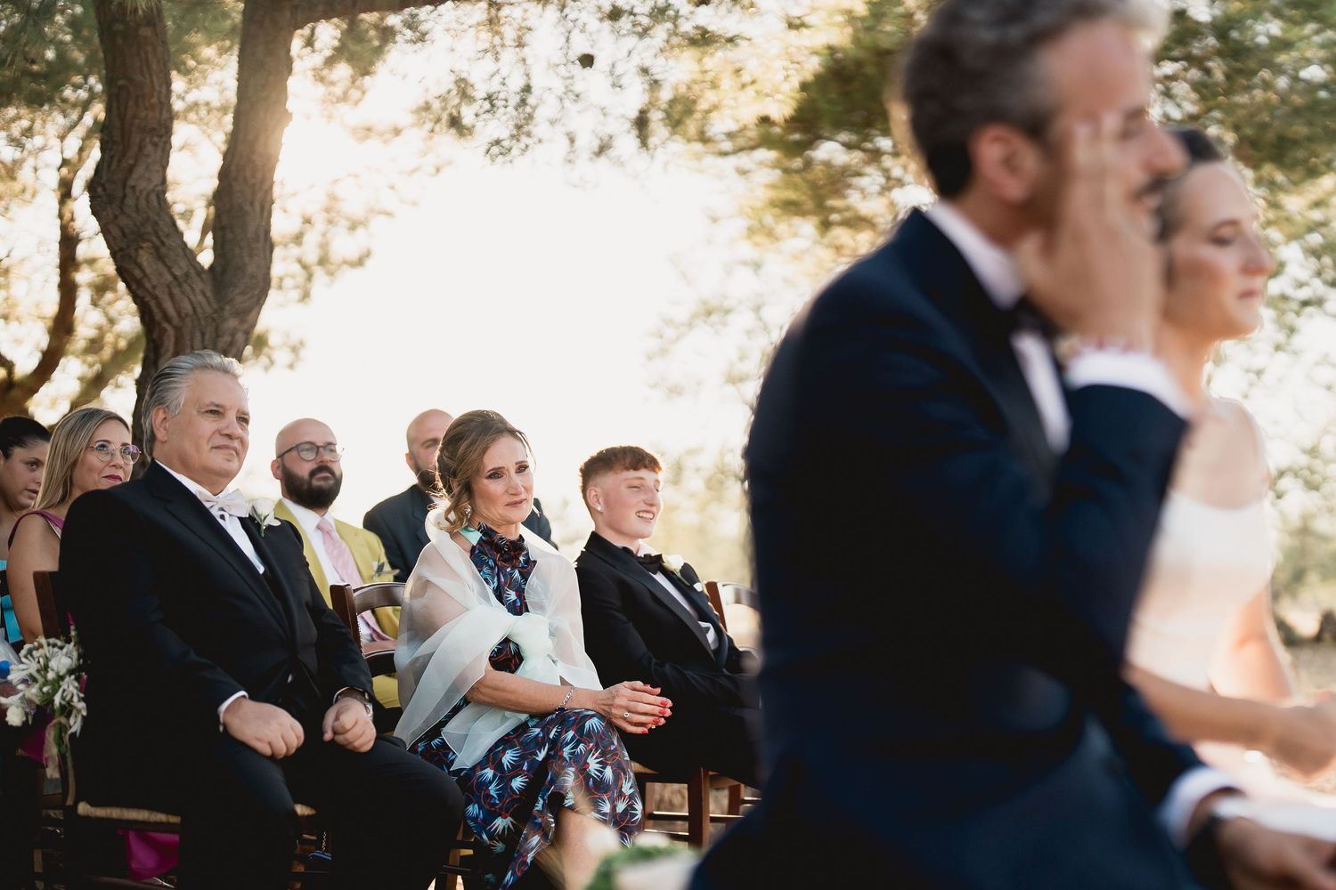 Wedding guests watching ceremony outdoors during golden hour with soft sunlit trees in background.