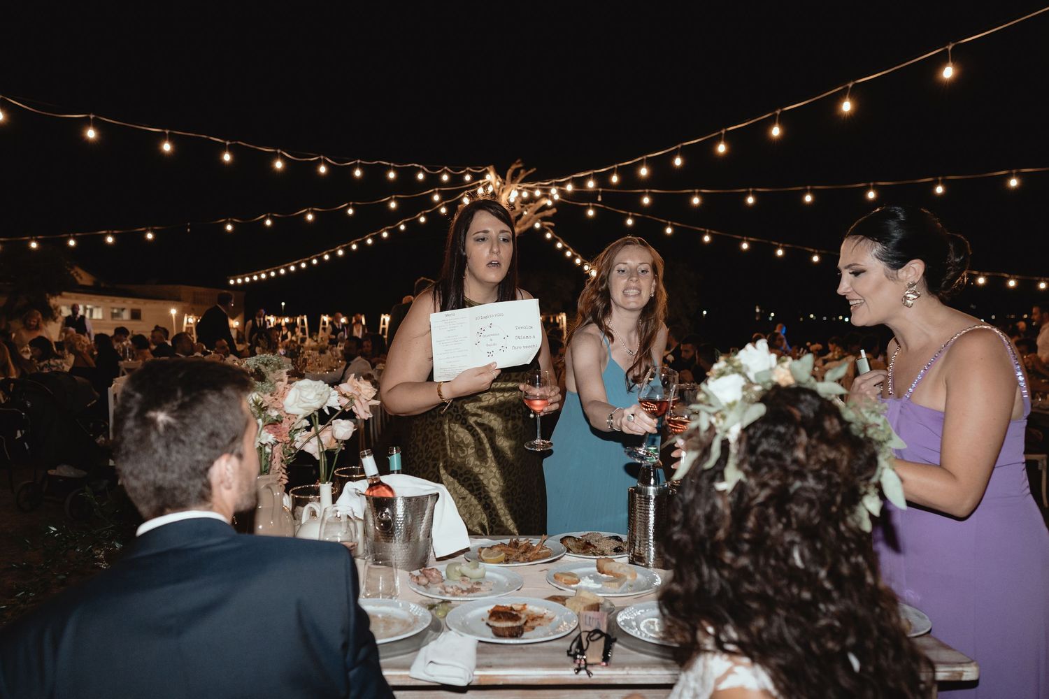 String lights illuminate an outdoor evening wedding reception with guests dining at long tables under the night sky.