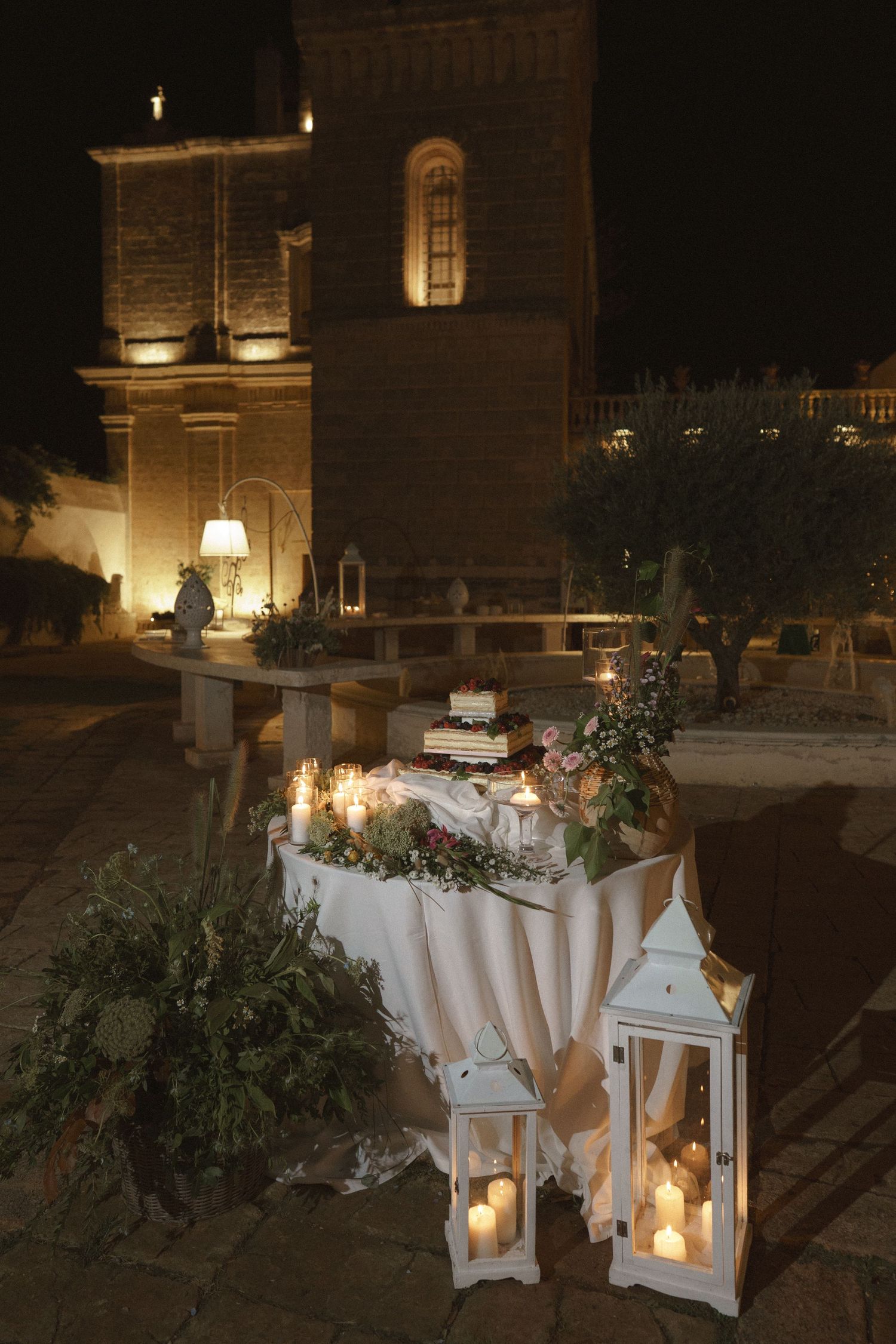 Elegant wedding decor with white lanterns and candles illuminated against a historic stone church at night.