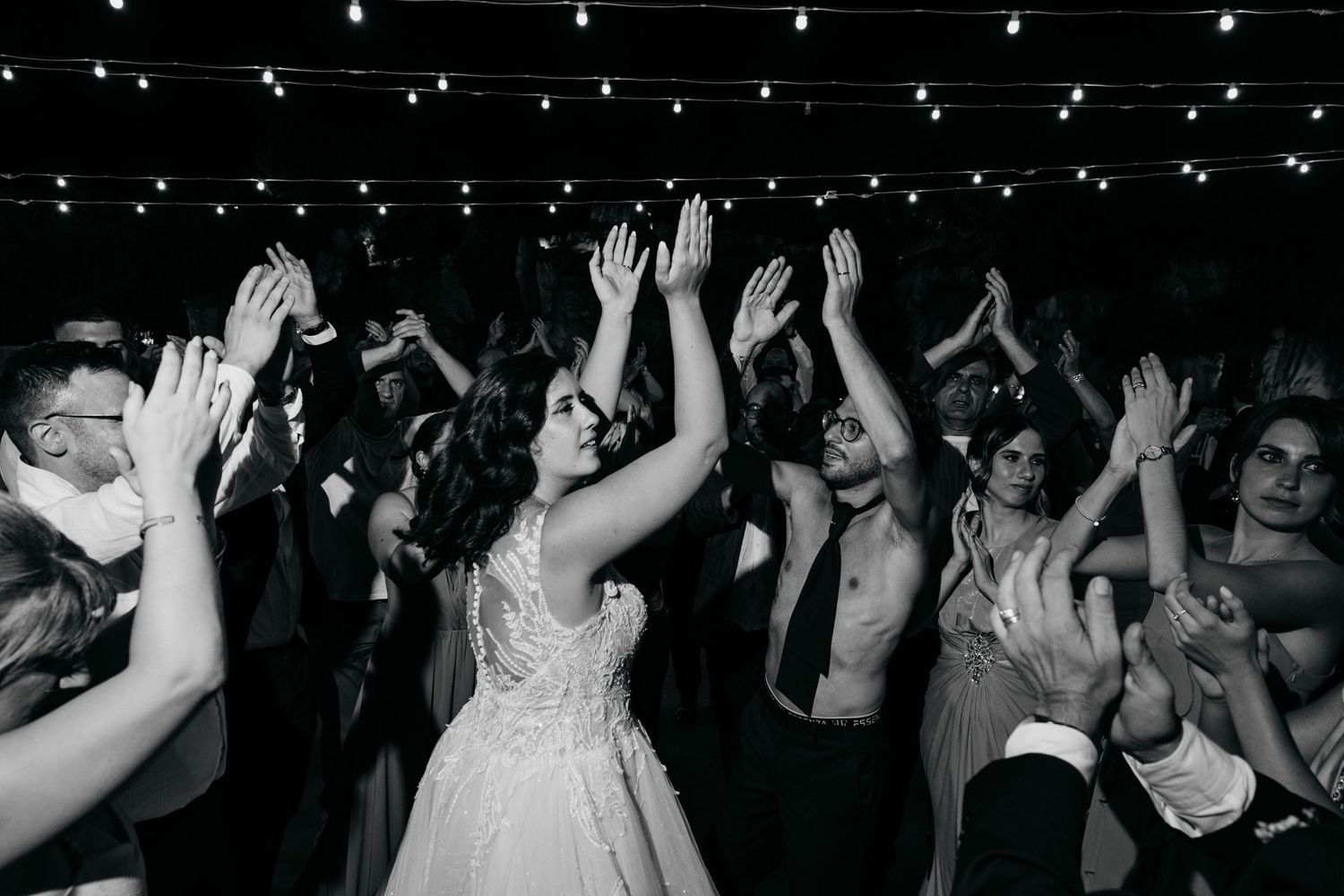 Wedding party guests dancing with raised arms under string lights at reception.