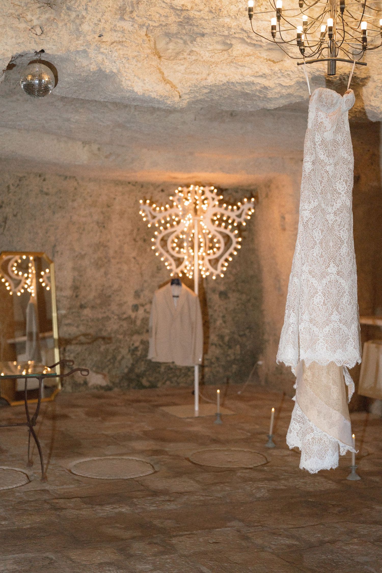 Lace wedding gown hanging beside a glowing chandelier and mirror inside a rustic stone cave venue with candles on the floor.