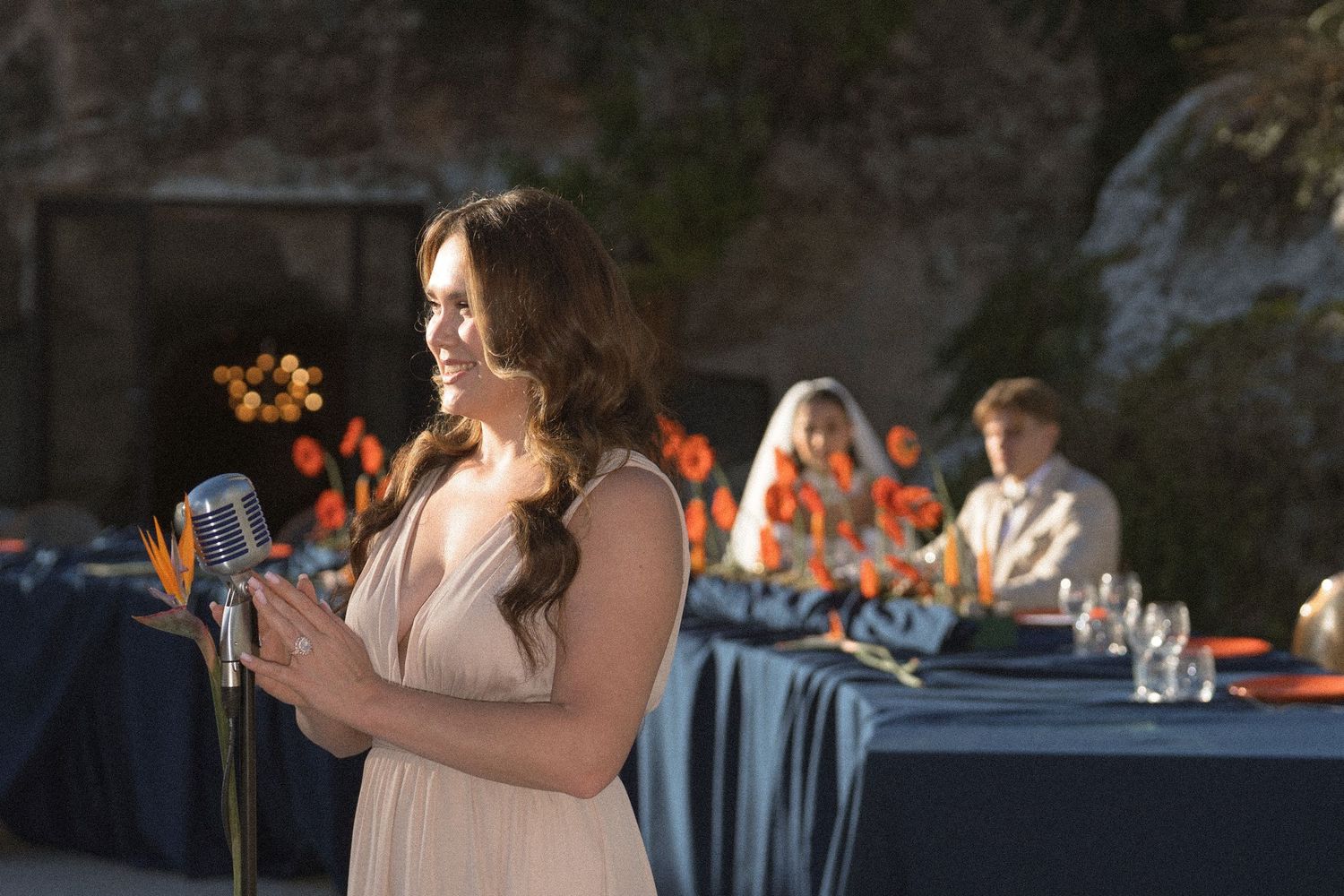 Woman in a blush dress sings into a vintage microphone at a wedding reception with orange flowers and guests seated at blue tables.