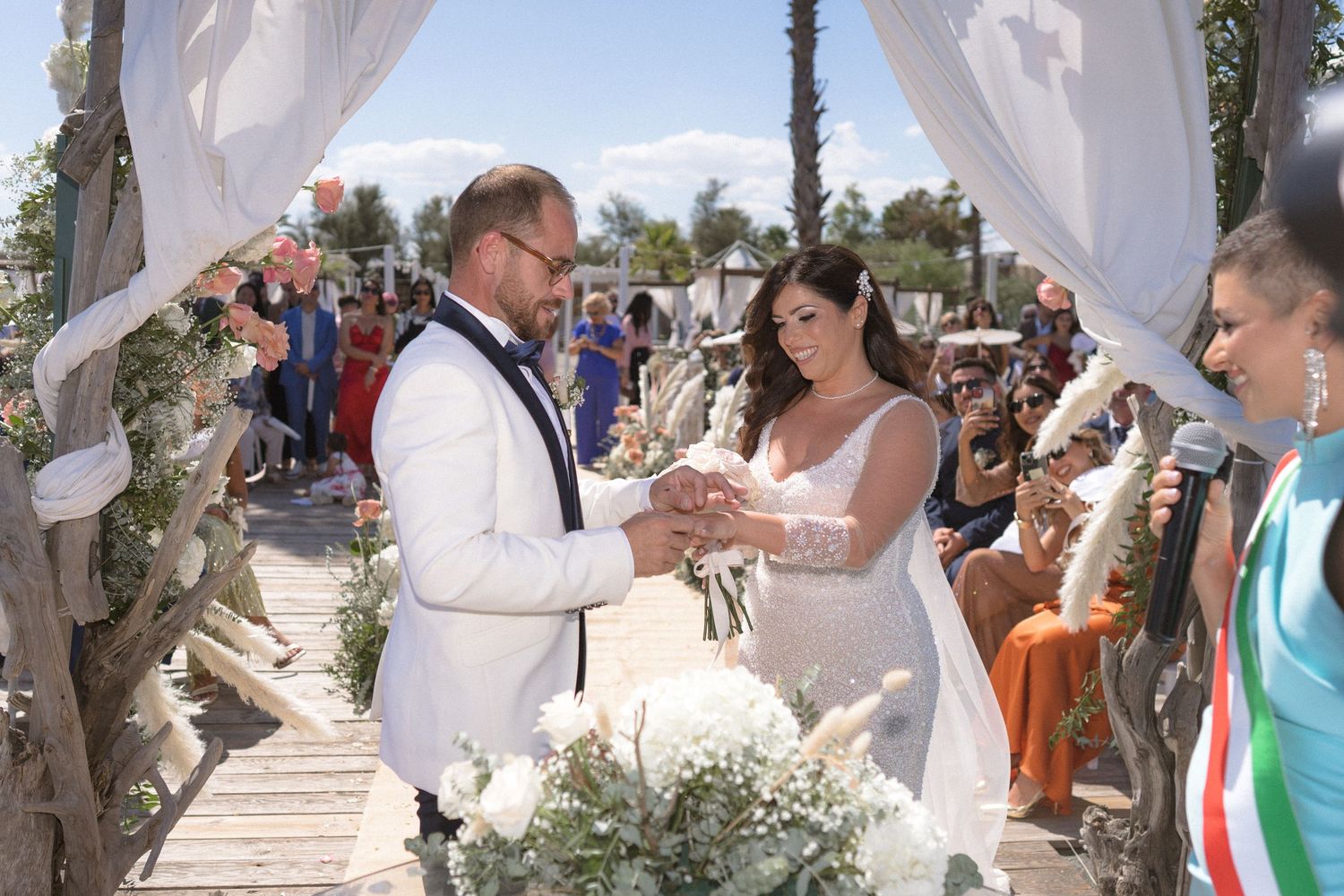 Wedding ceremony taking place under a white draped canopy with guests seated outdoors.