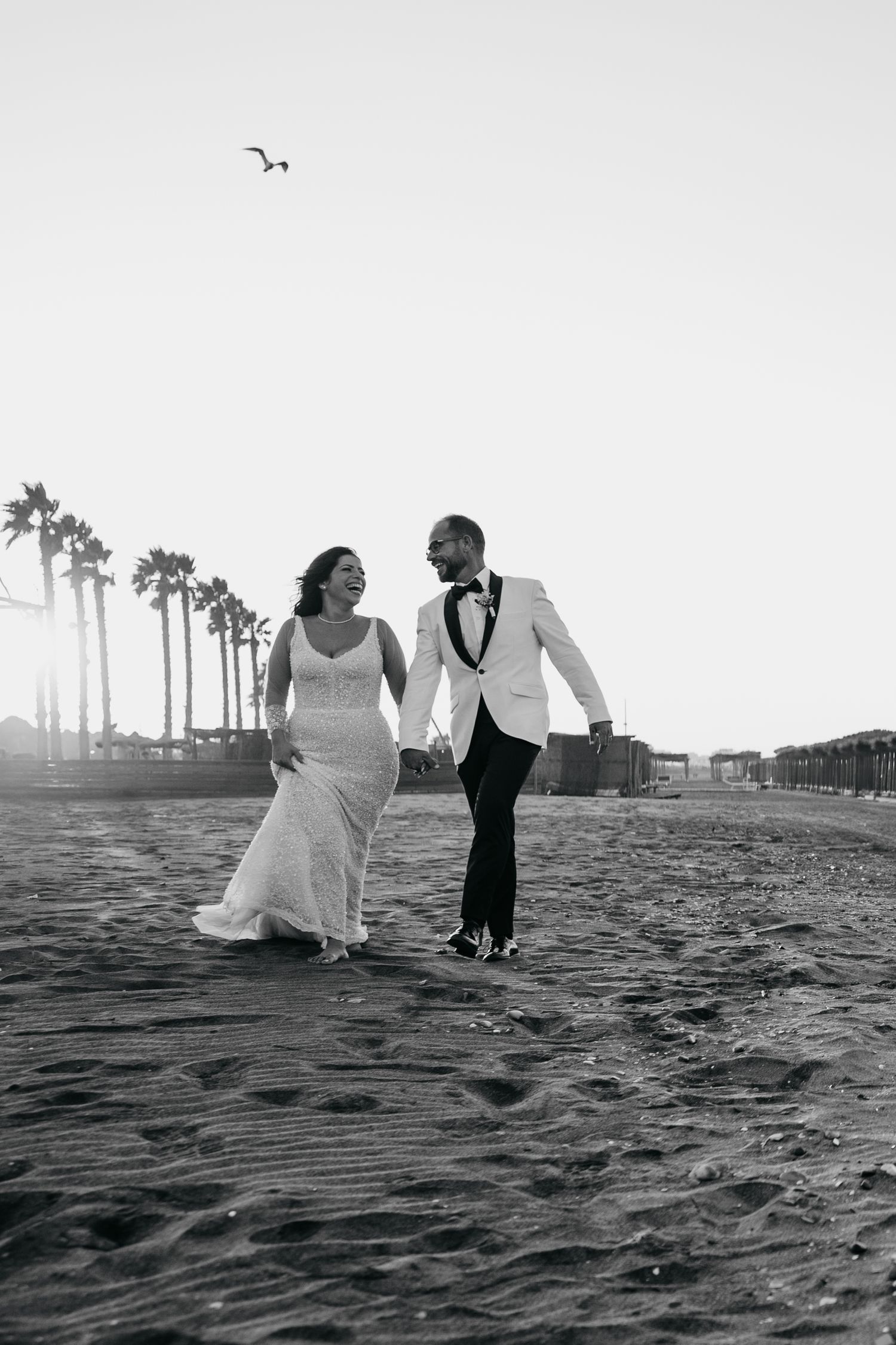A couple in wedding attire strolling on a beach with palm trees and seabirds in the background.