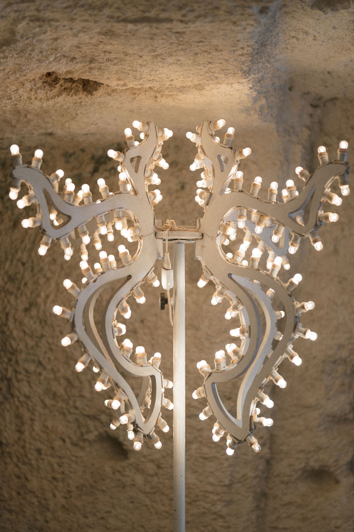 White branching floor lamp with symmetrical antler-like arms covered in small bulbs, set against a rough textured stone wall.