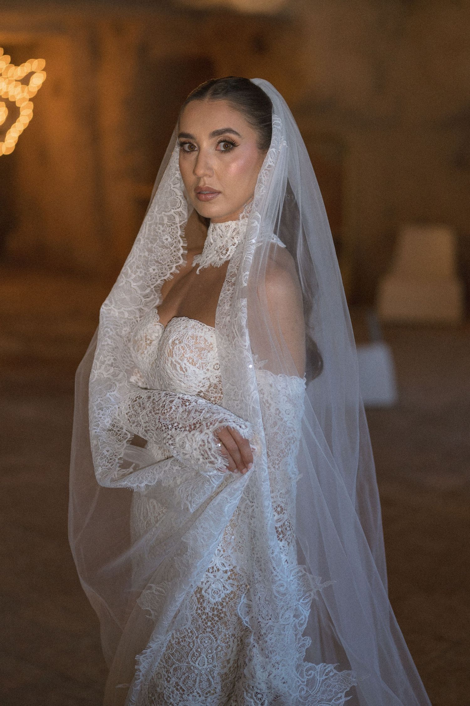 Bride wearing a lace-trimmed cathedral veil and strapless lace gown with sheer sleeves stands in a softly lit stone venue.
