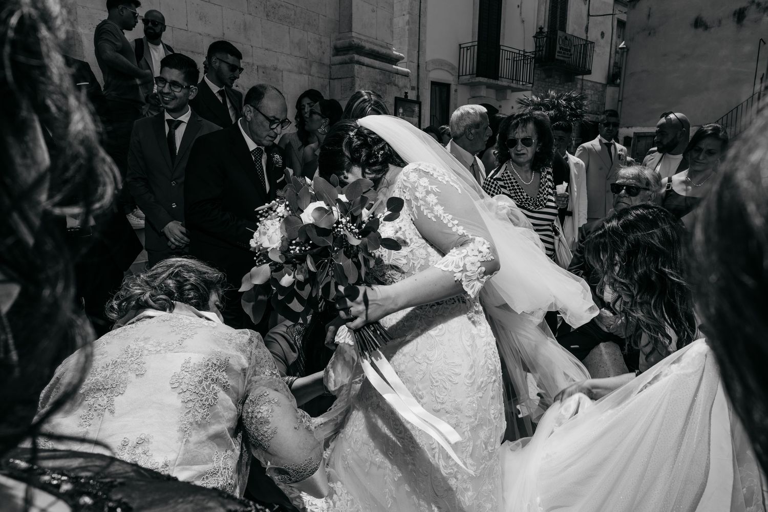 Black and white wedding photo of bride in white gown holding bouquet surrounded by guests outside a church.