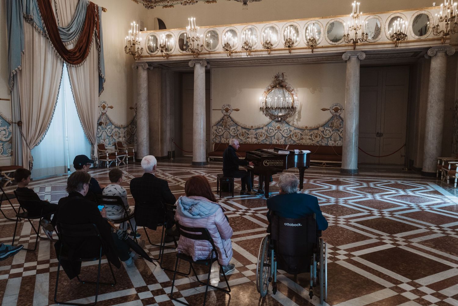 Classical music performance in an ornate palace room with decorative moldings and chandeliers.