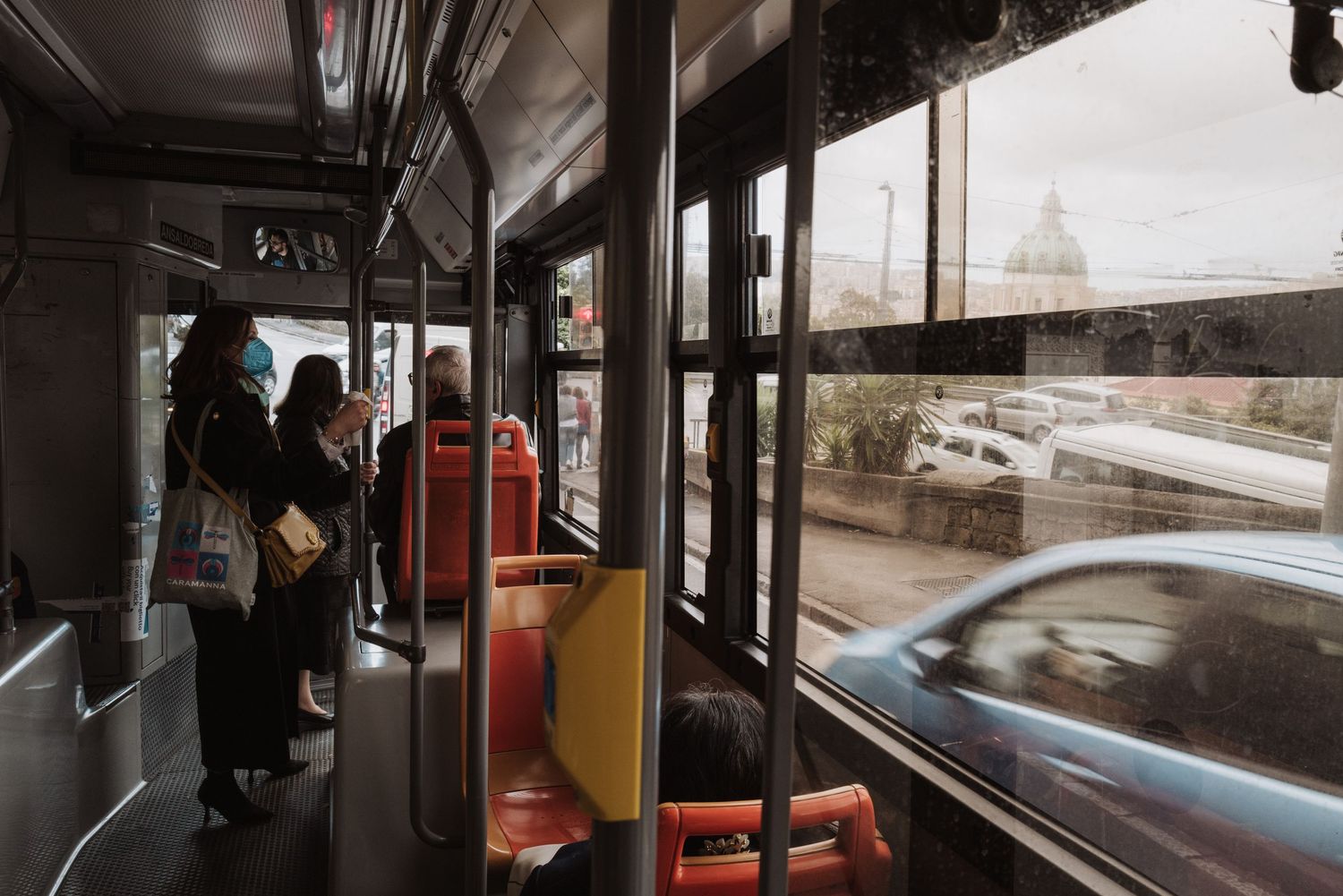 A passenger rides on a public transit bus with a view of city streets and passing cars through the window.