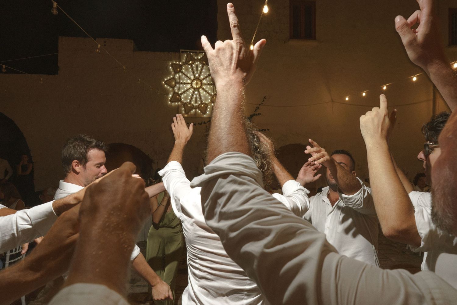 Enthusiastic wedding guests dancing with hands raised under a decorative ceiling light.