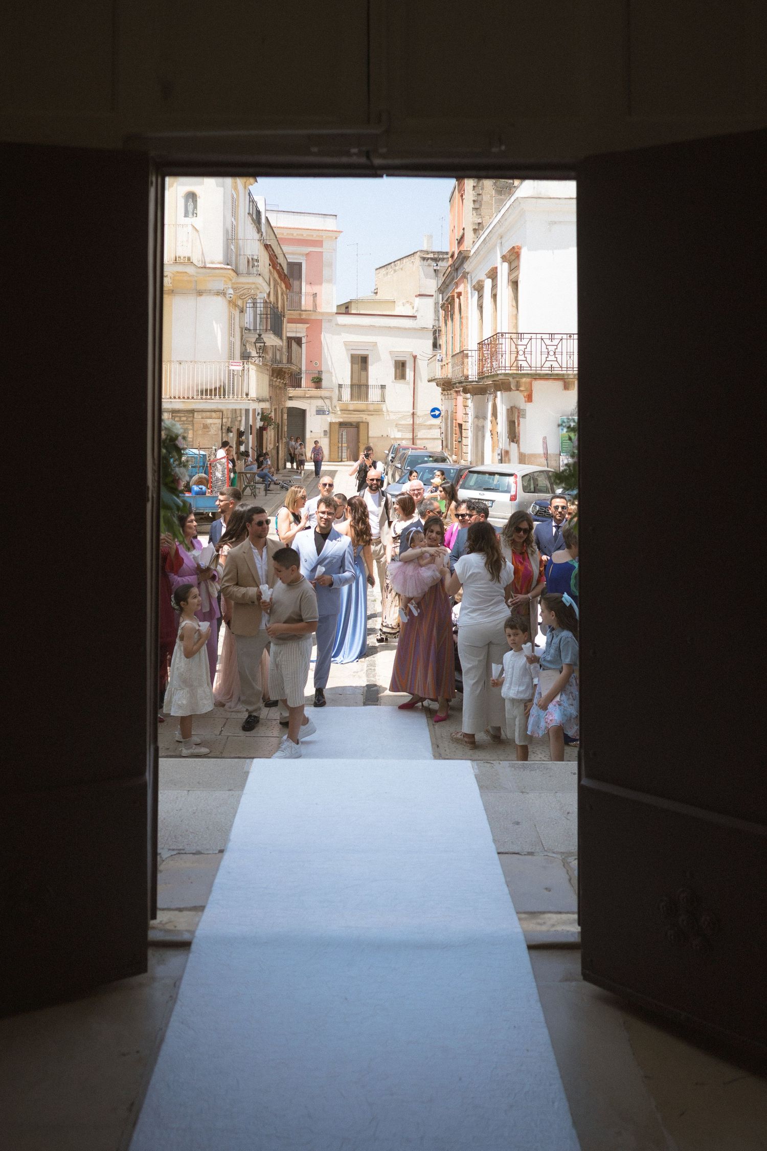 People gather under archways in a shaded corridor looking out onto a sunlit street in a Mediterranean town.