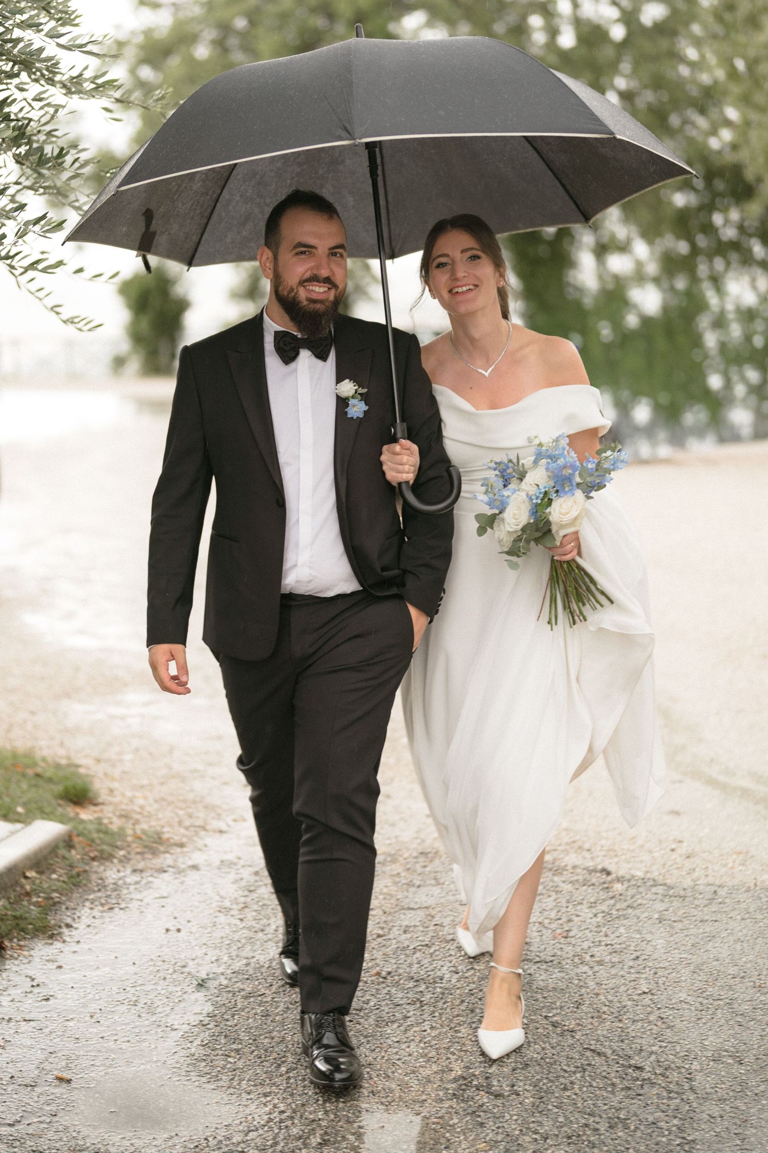 Couple walking together under black umbrellas on a rainy gravel path during their wedding day.