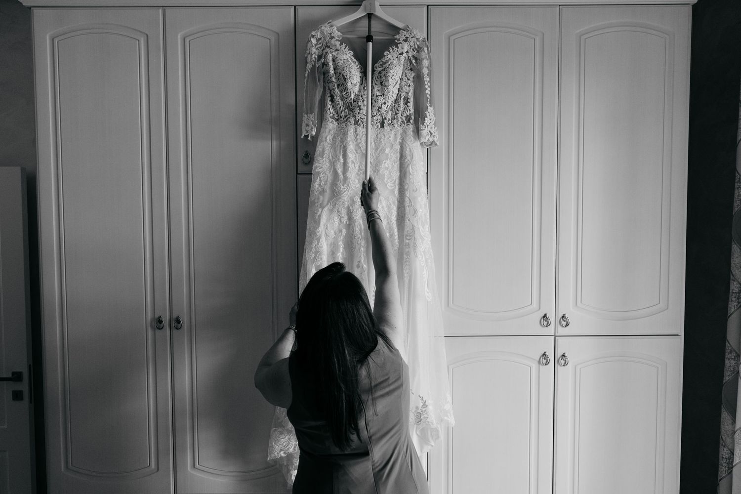 Black and white photo of someone reaching up to touch a white dress hanging on a wardrobe.