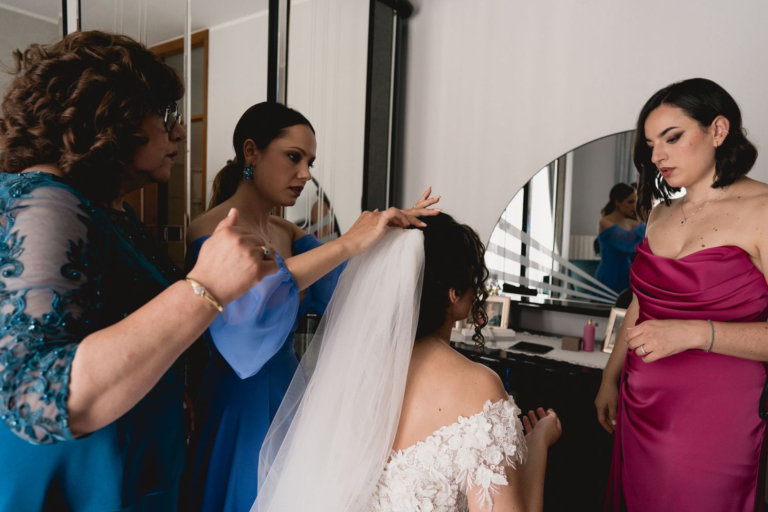 Wedding preparation scene with attendants in blue and burgundy dresses helping with final touches.