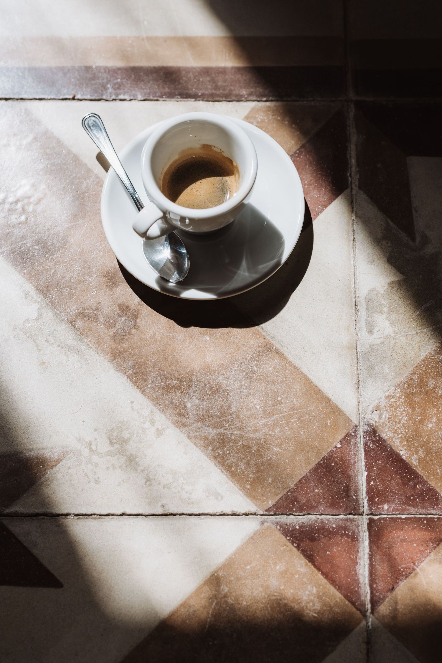 Empty espresso cup on saucer casting dramatic shadows on geometric tiled floor.