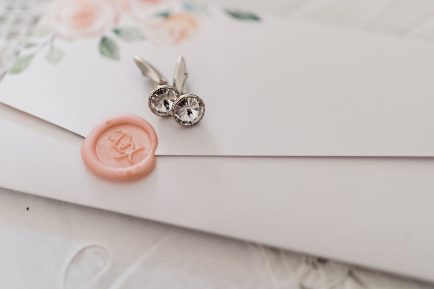 Wedding invitation with pink wax seal and silver earrings on white surface.