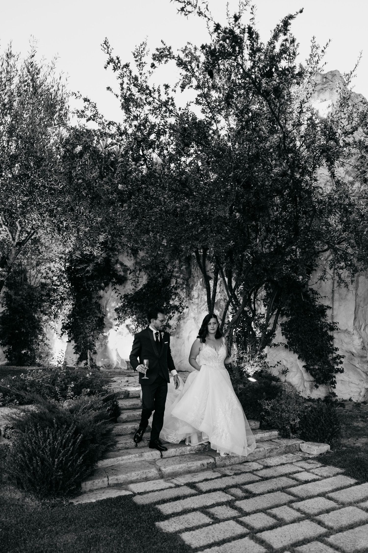 A black and white wedding photo of a couple walking through a lush garden archway on stone pavers.