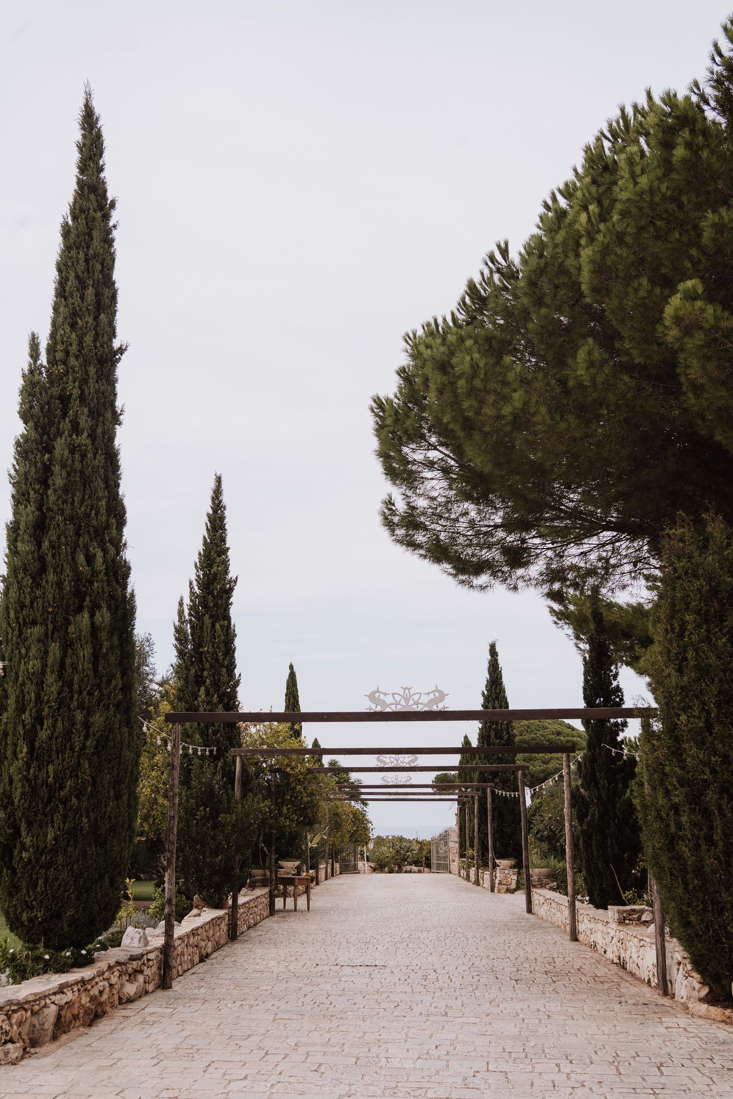 A stone pathway lined with tall cypress trees and wooden pergola creates a Mediterranean atmosphere.