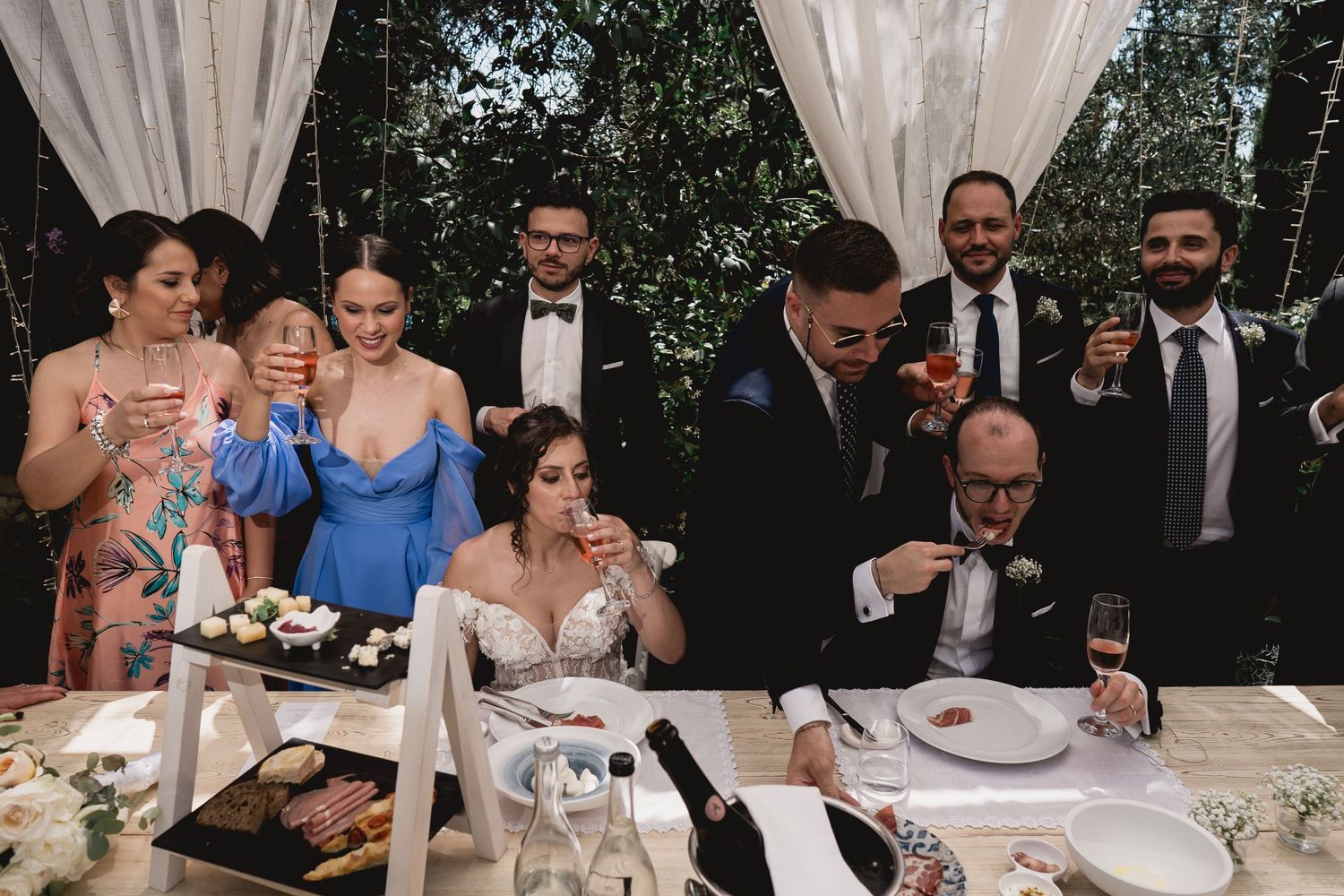 Wedding guests enjoying a lively reception dinner outdoors under white drapes with trees in the background.
