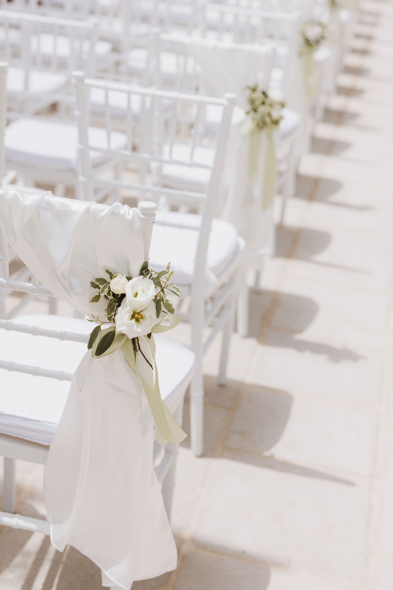 White wedding ceremony chairs decorated with floral arrangements and ribbon.