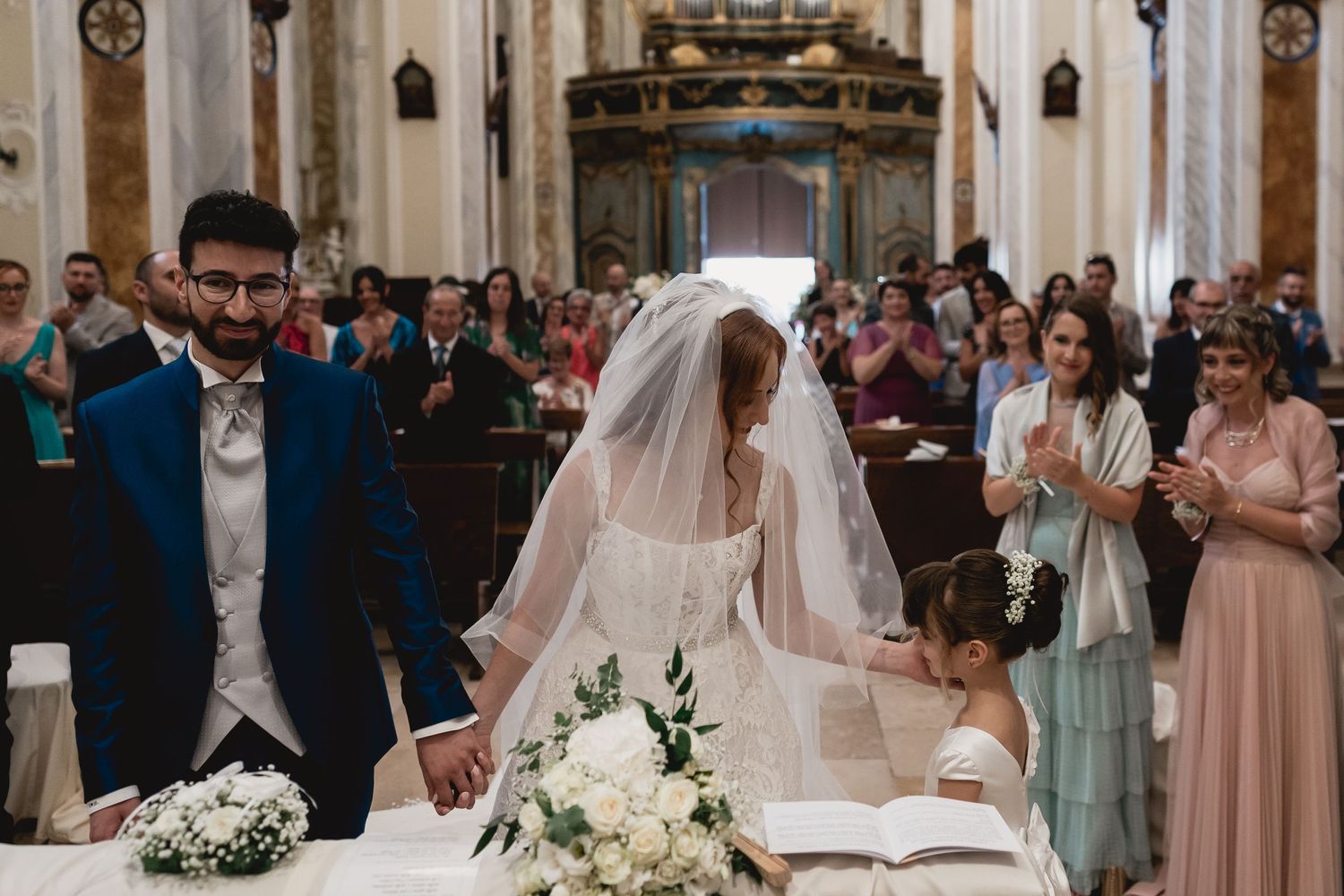 Wedding ceremony in grand church with couple at altar and guests in pastel dresses.