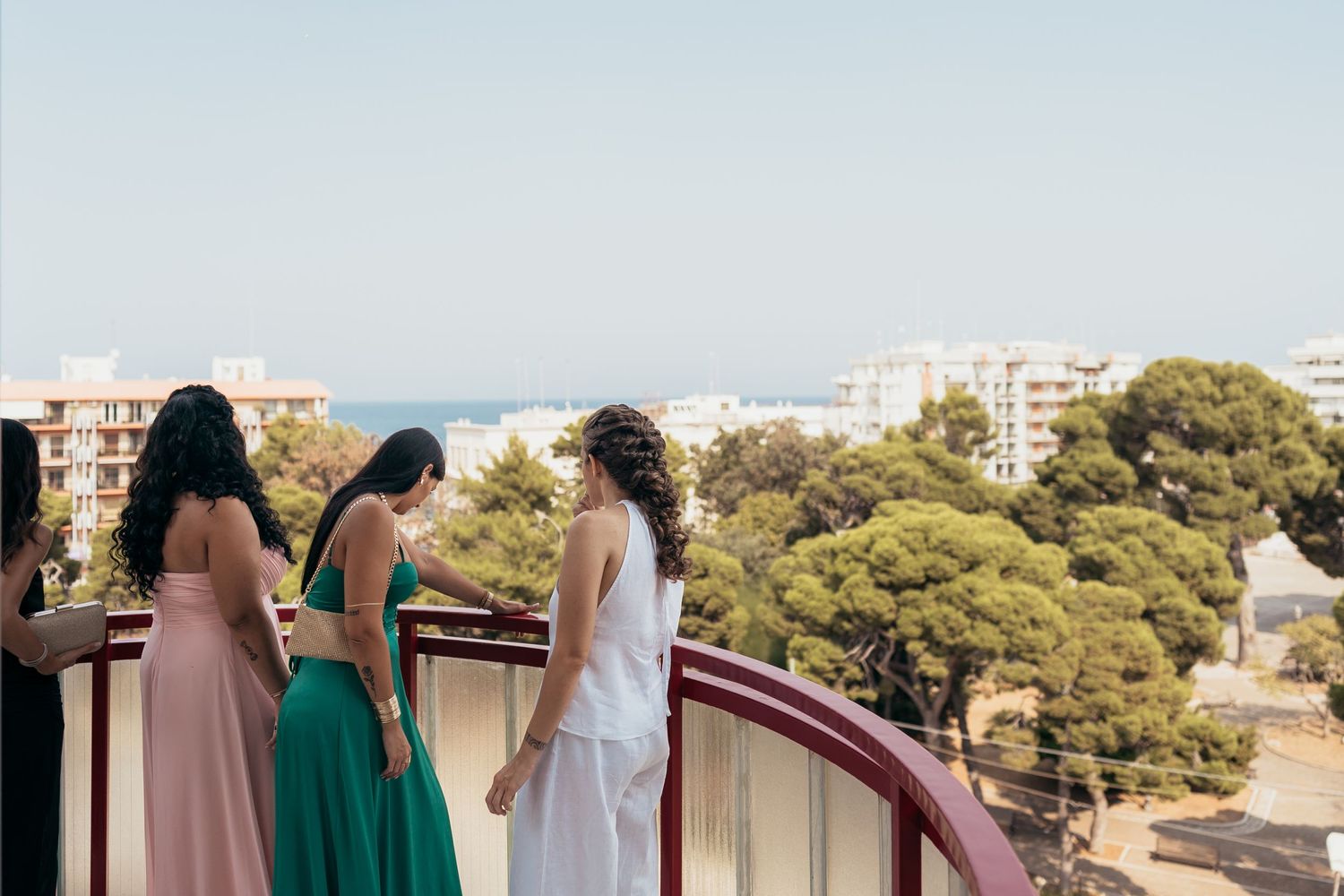 Three people in formal dresses stand on a balcony overlooking a coastal cityscape.
