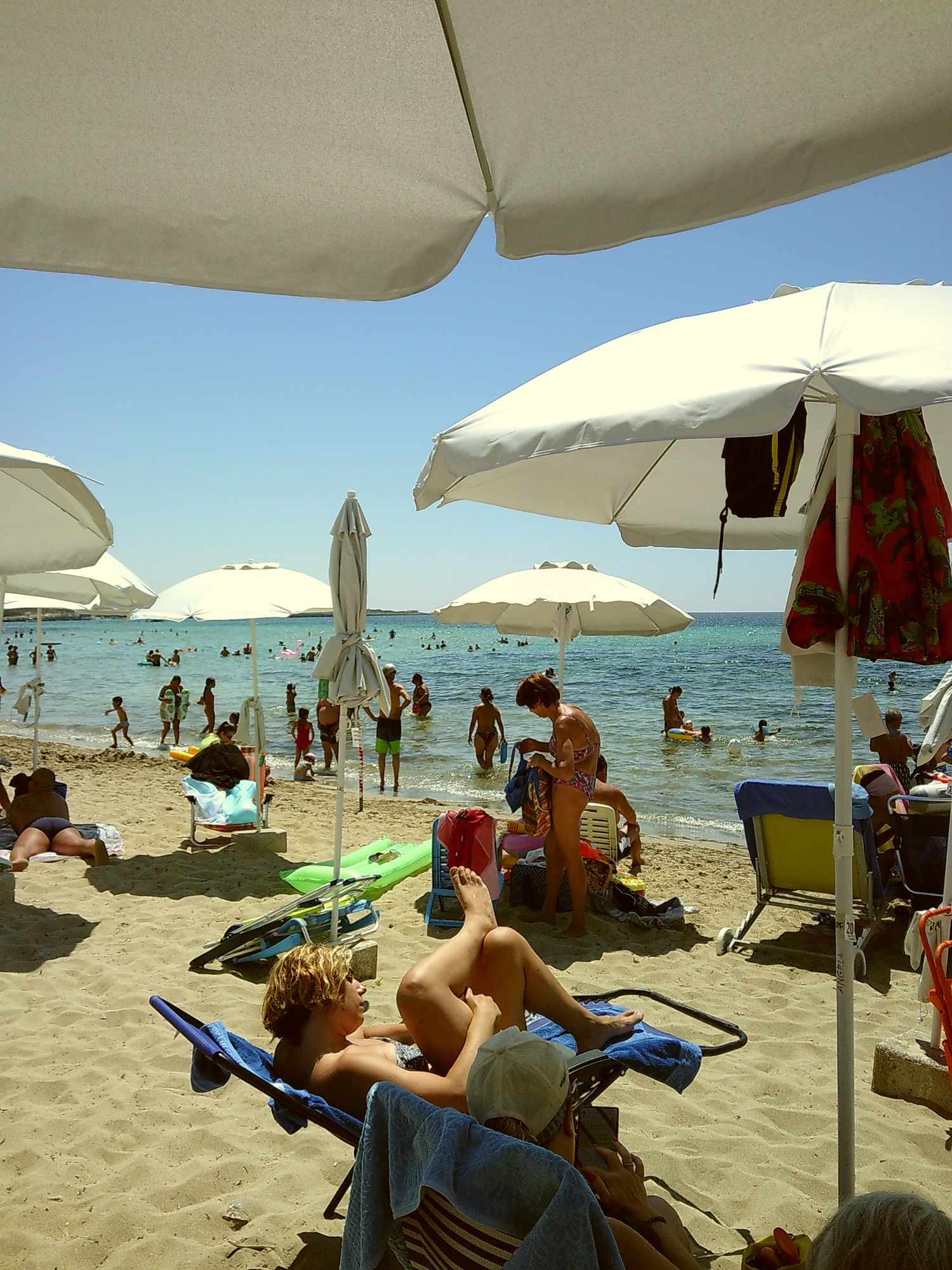 Crowded beach scene with white umbrellas, beachgoers, and crystal blue waters on a sunny day.