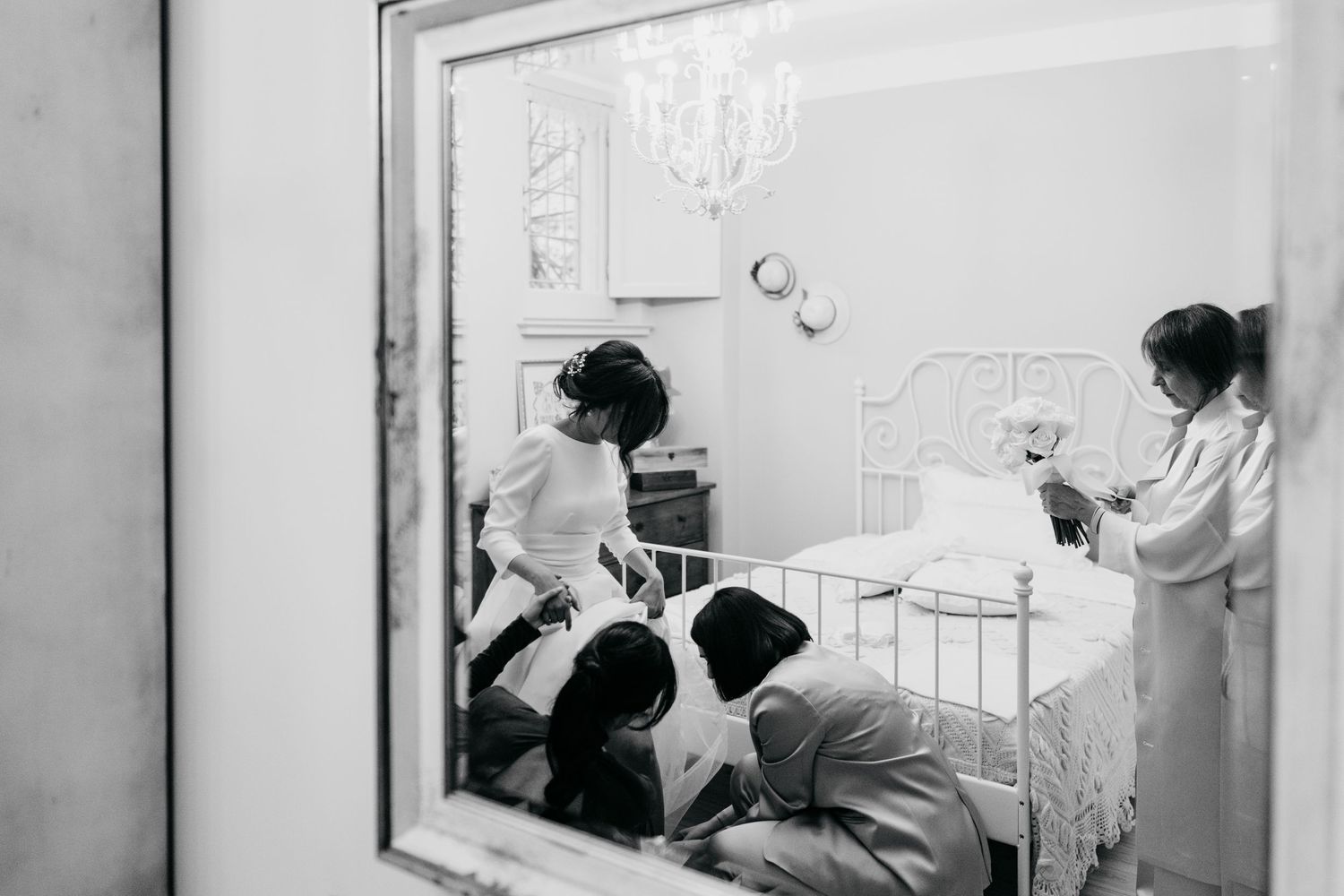 Black and white mirror reflection of bridesmaids helping a bride prepare in an elegant white bedroom.