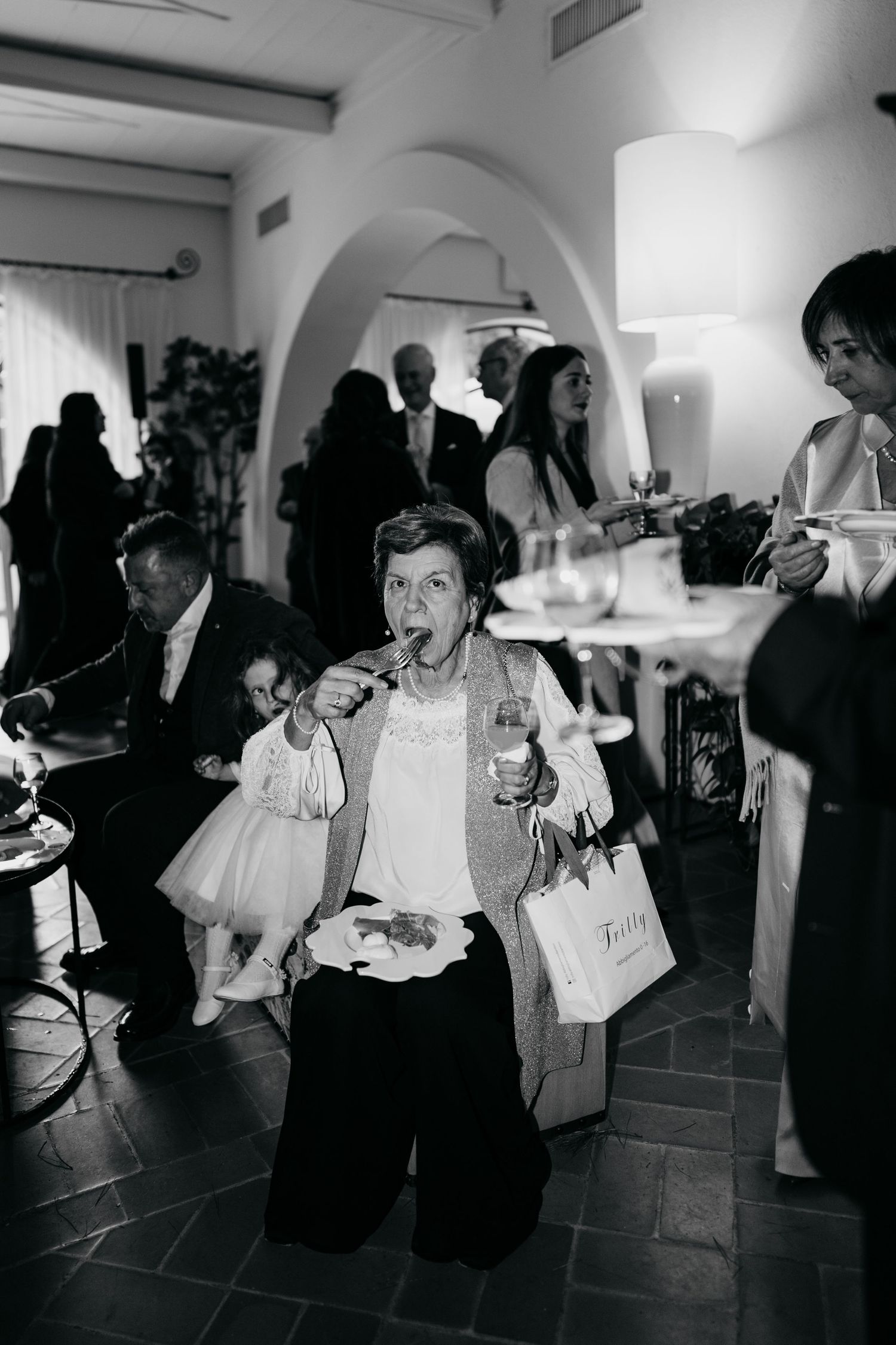 Elderly woman eating and holding a wine glass at a wedding reception in black and white photo.