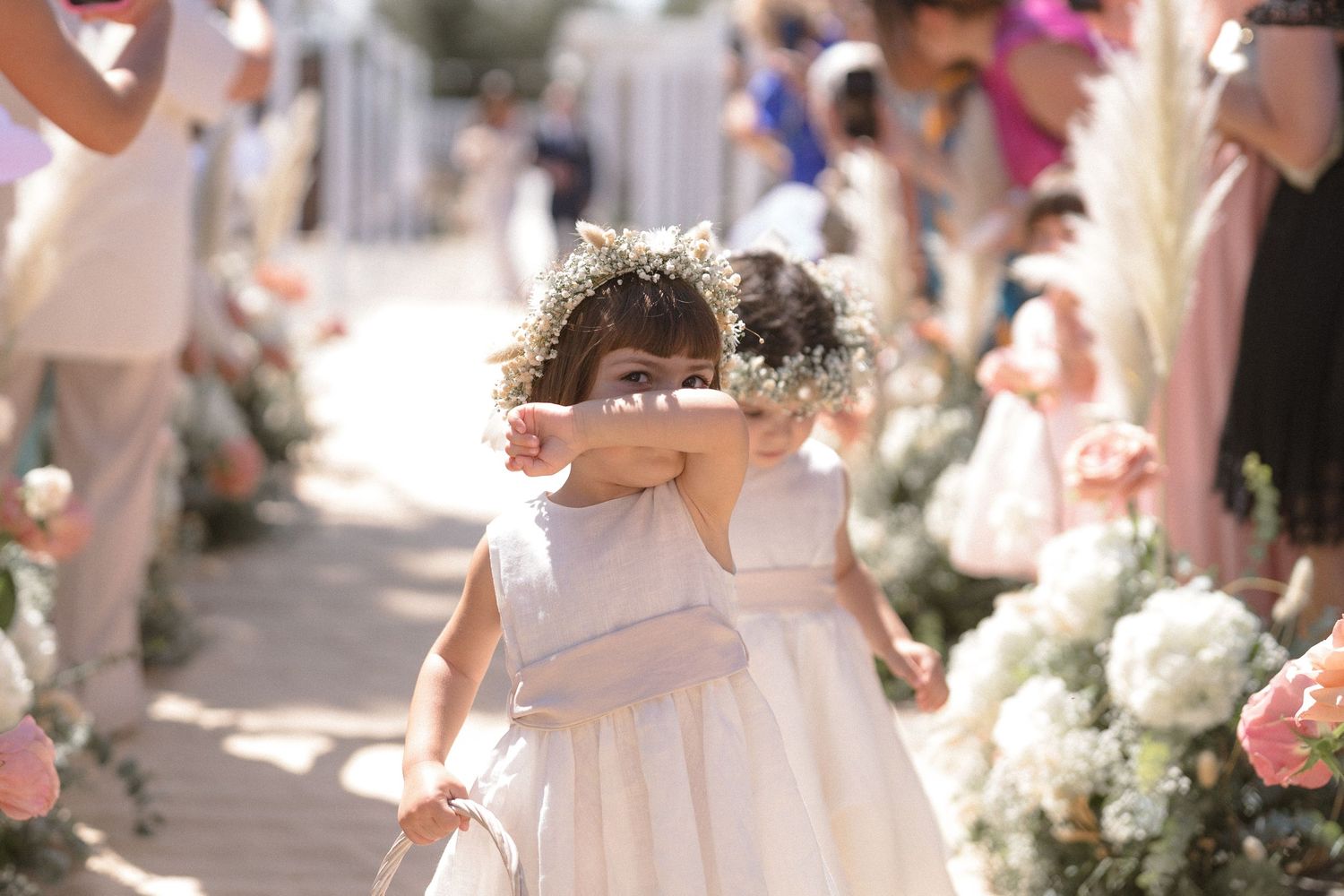Young flower girls in white dresses walk down an aisle decorated with pink and white flowers.