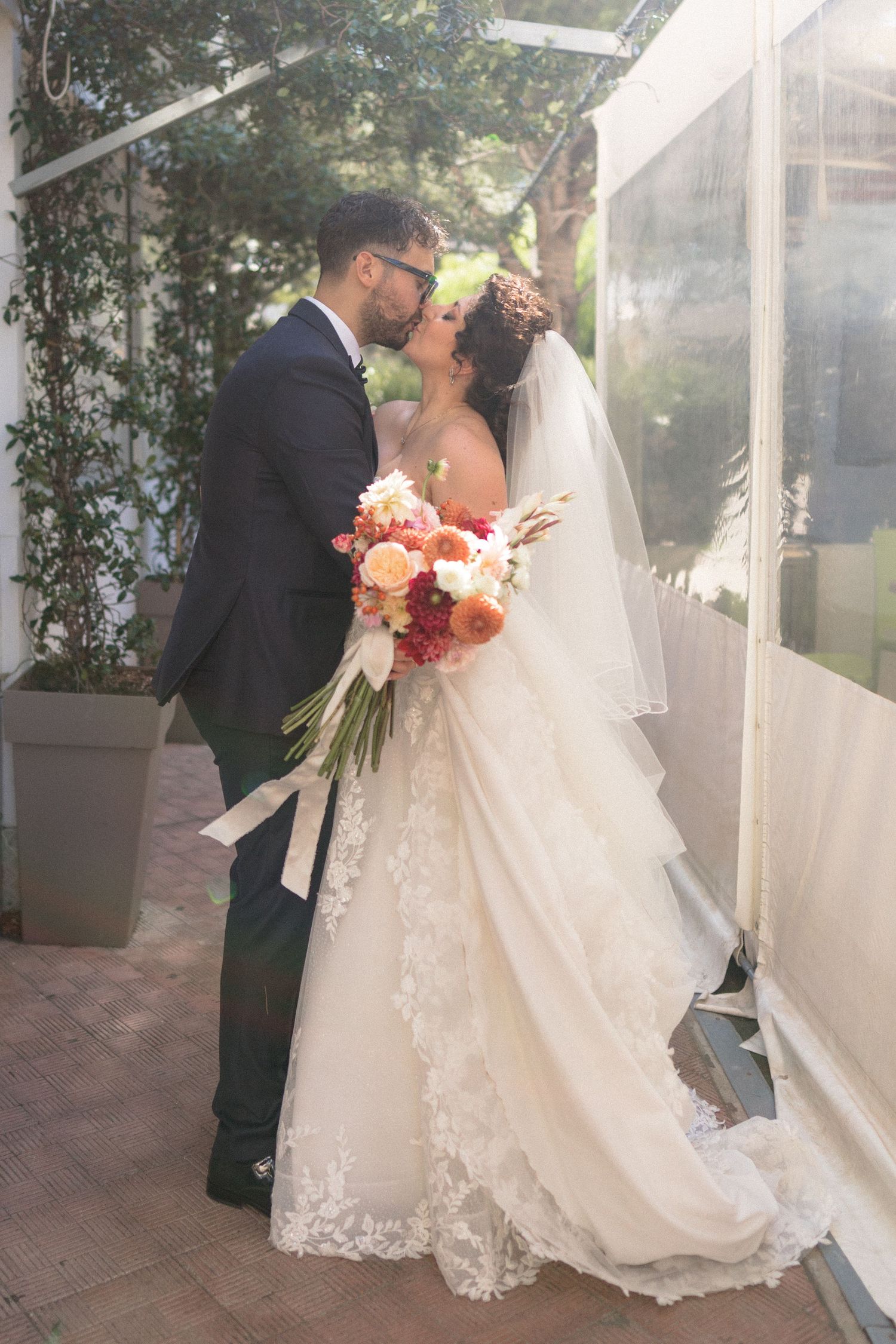 Bride and groom sharing a romantic kiss, bride holding a vibrant bouquet of coral and pink flowers.