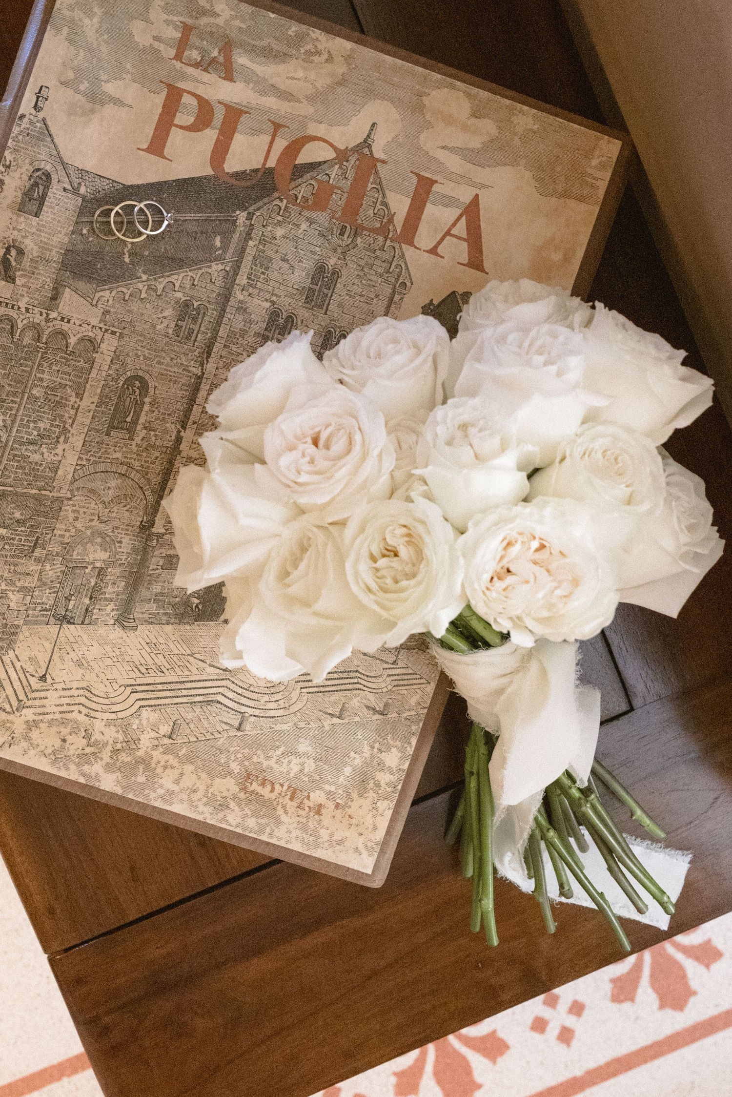 White garden rose bridal bouquet beside a vintage La Puglia book with wedding rings on a wooden surface.