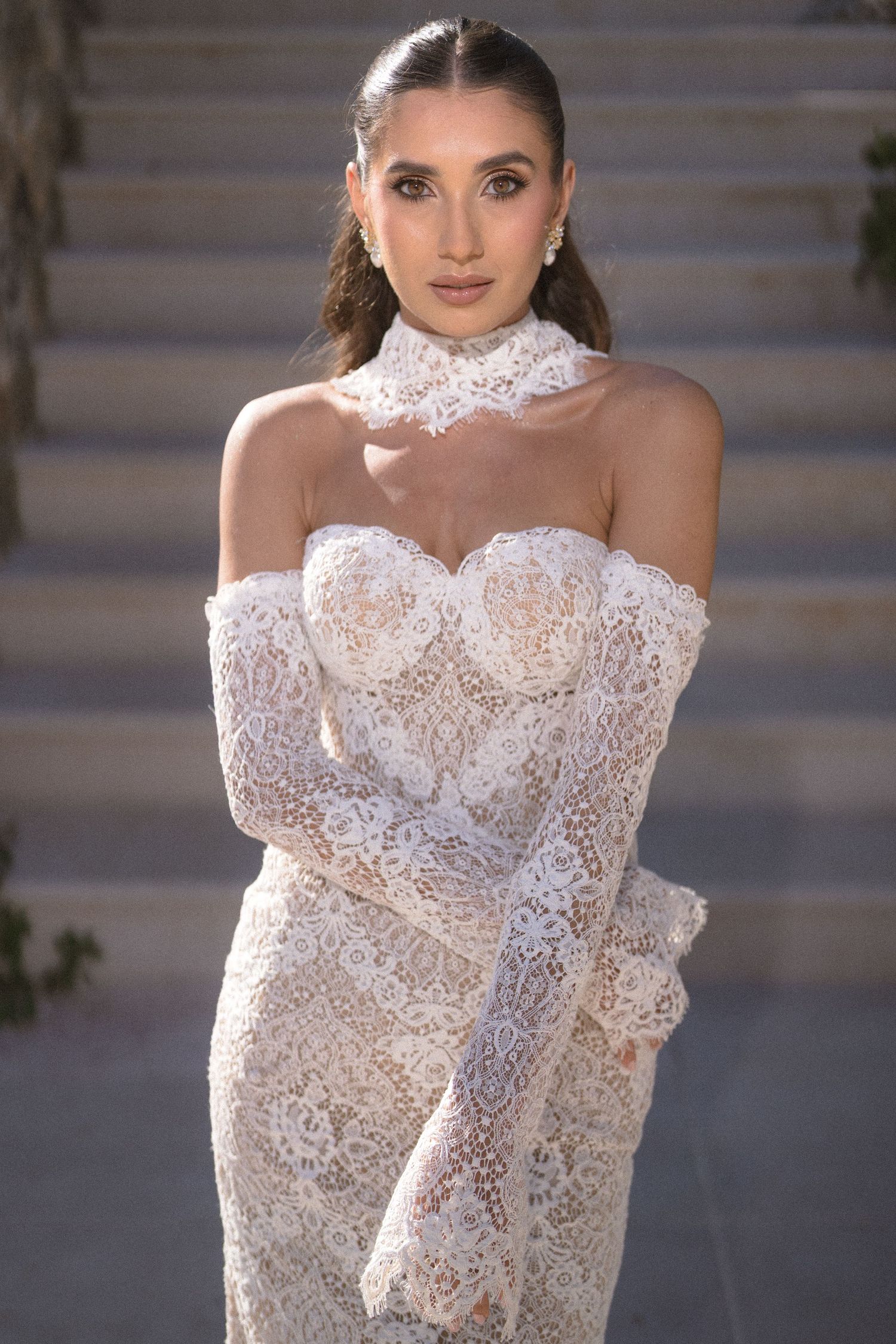 Close-up portrait of a bride in a strapless lace wedding gown with a lace choker, standing before sunlit stone steps.