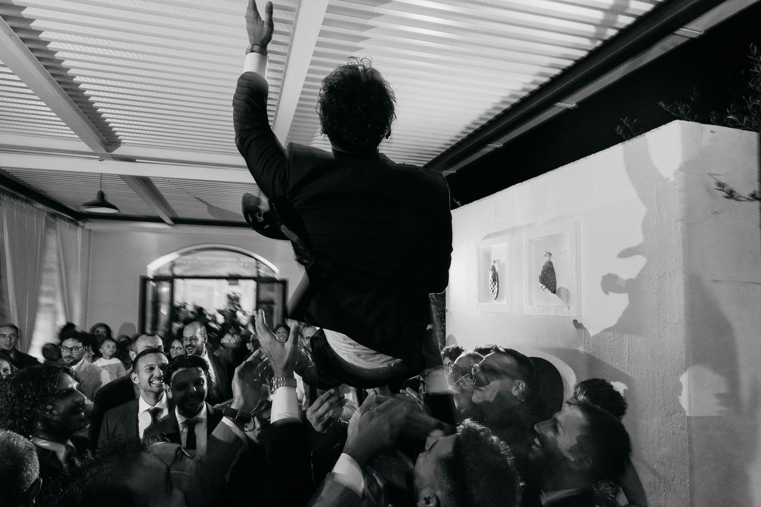 Groom crowd surfs with arm raised above celebrating wedding guests in black and white photo.