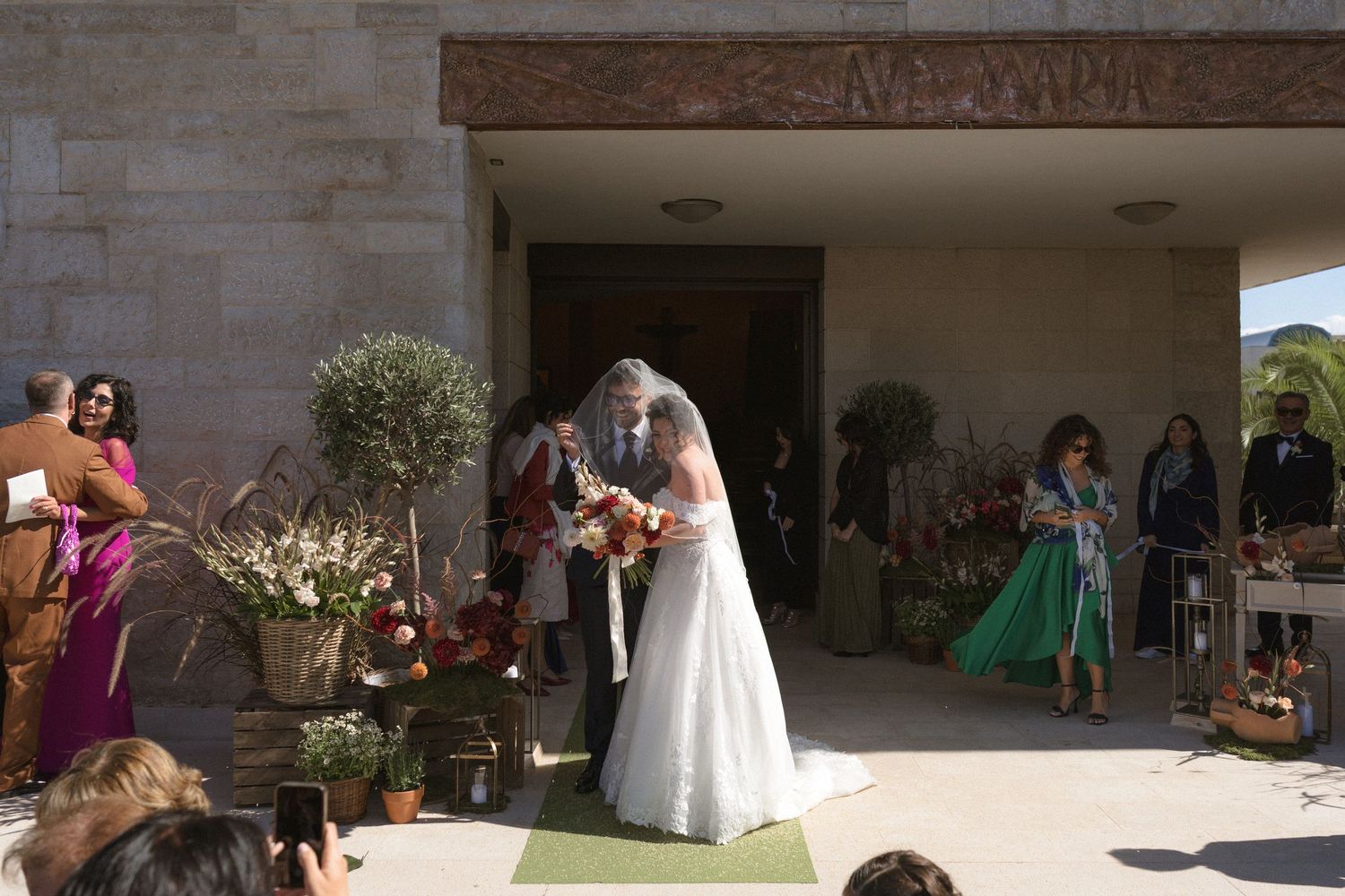 Bride and escort share embrace at church entrance surrounded by floral decorations and guests.