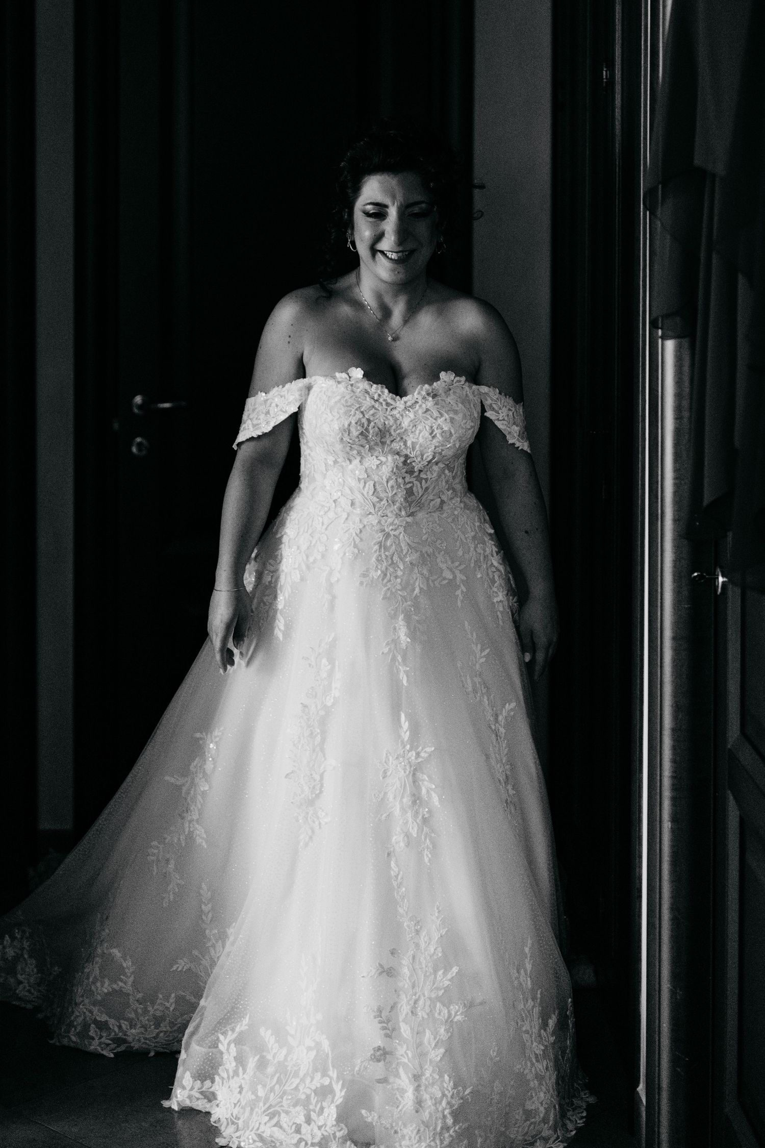 Smiling bride in off-shoulder lace ball gown wedding dress poses in doorway in black and white photo.