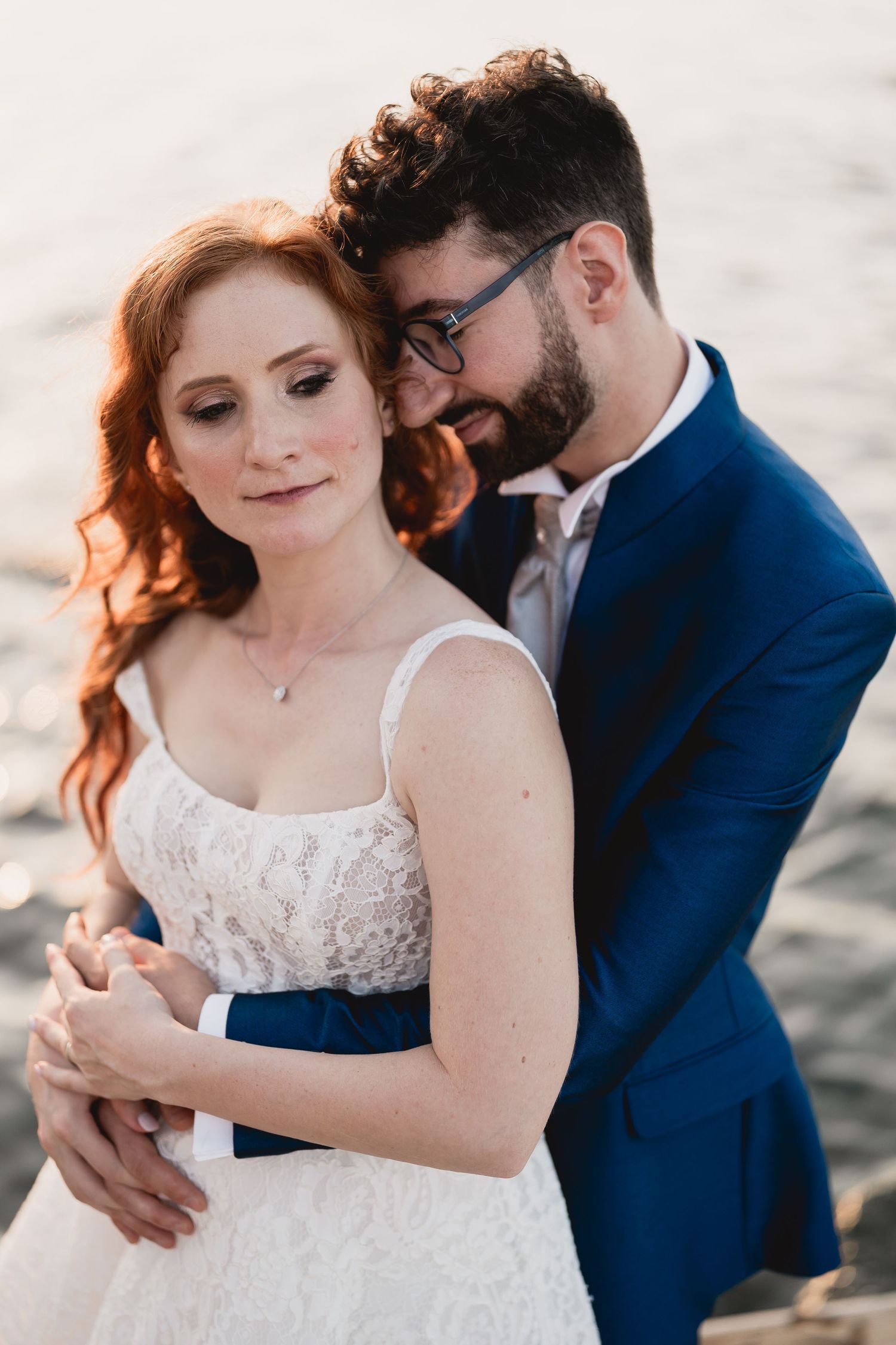 Romantic couple embracing at sunset, with bride in lace dress and groom in navy suit.