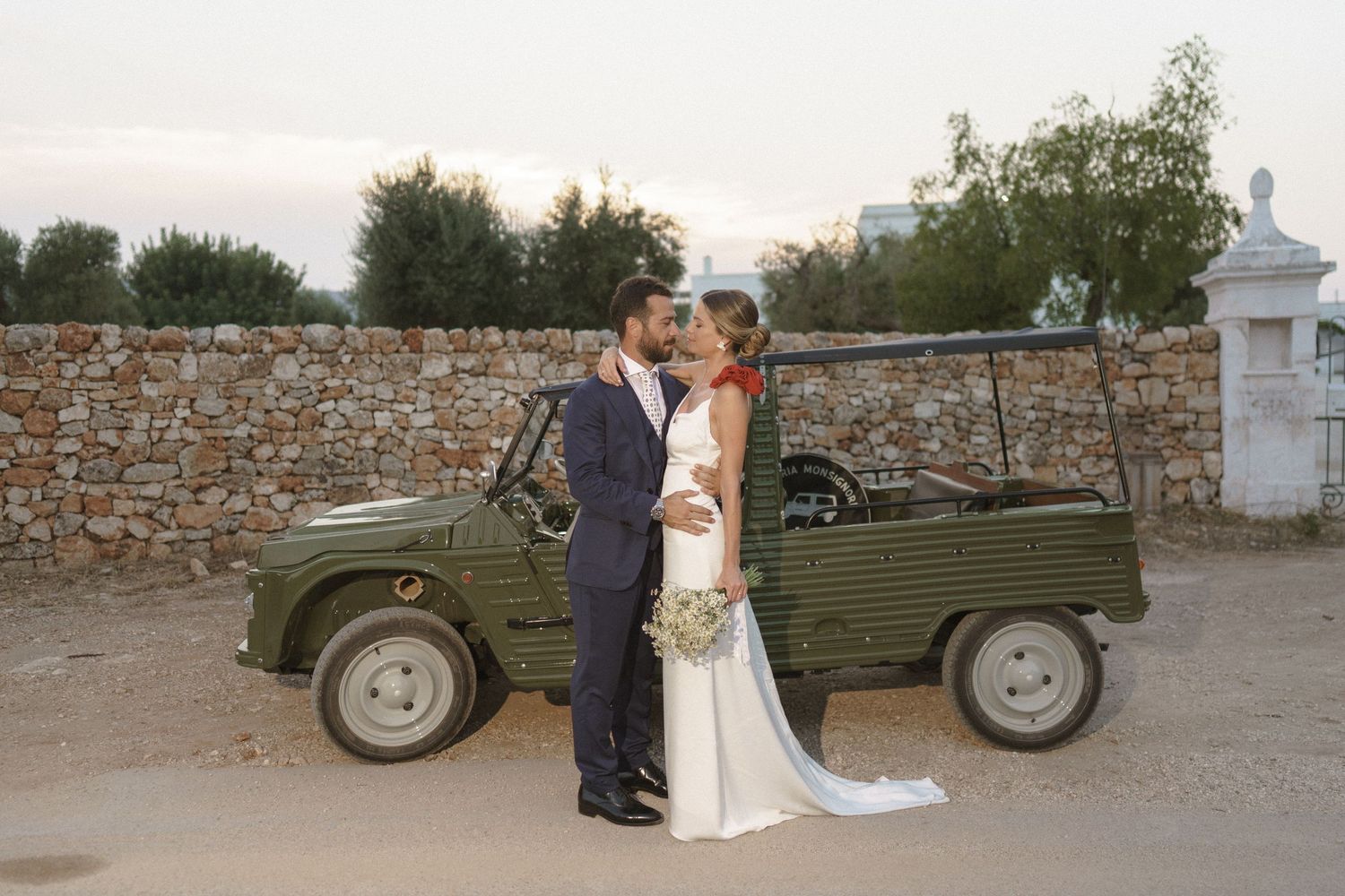 Vintage green military jeep serves as wedding photo backdrop with couple posing against stone wall.