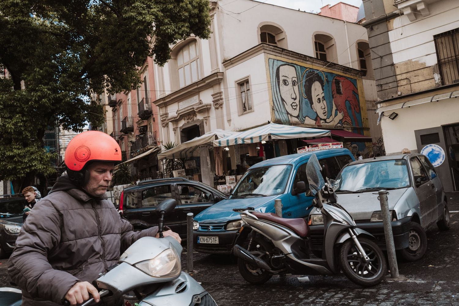 A motorcyclist wearing a red helmet and winter coat rides through a city street with parked cars and murals on buildings.
