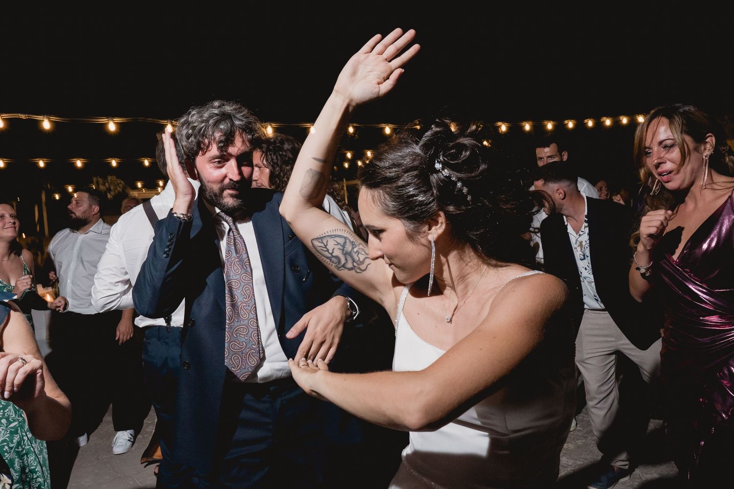 People dancing and celebrating at a lively outdoor wedding reception with string lights overhead.