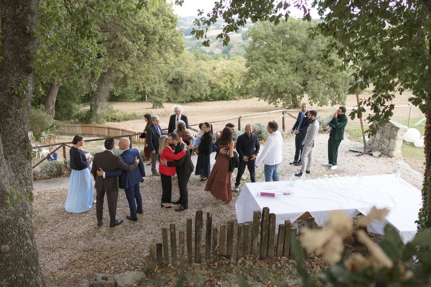 Group gathering at an outdoor event with tables set up under shady trees in a natural setting.