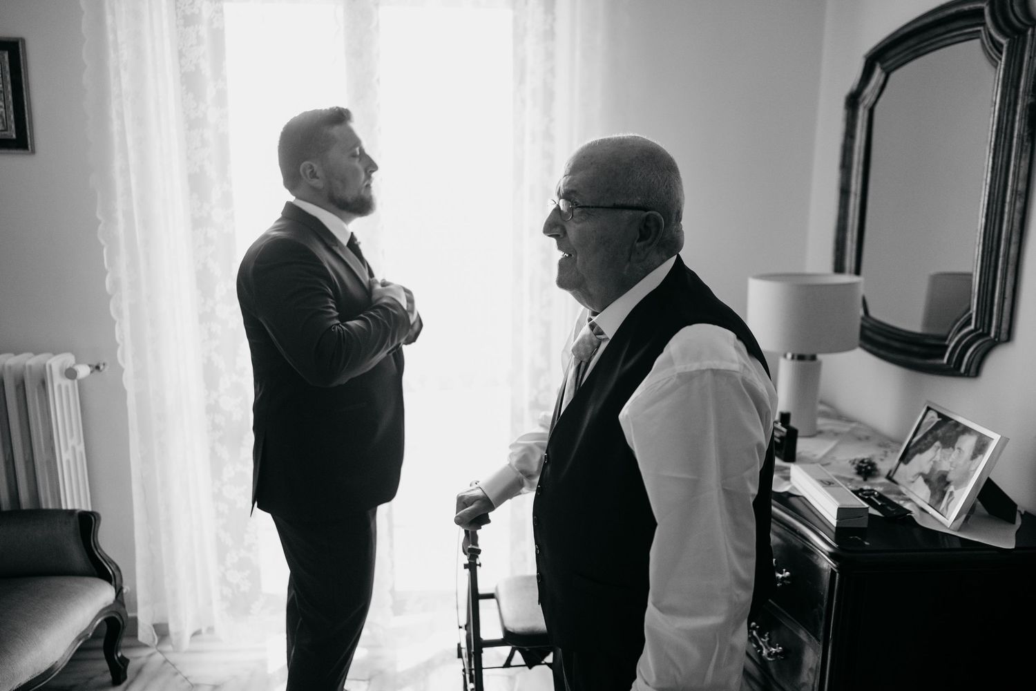Two elderly men in formal attire converse by a window in a black and white photograph.