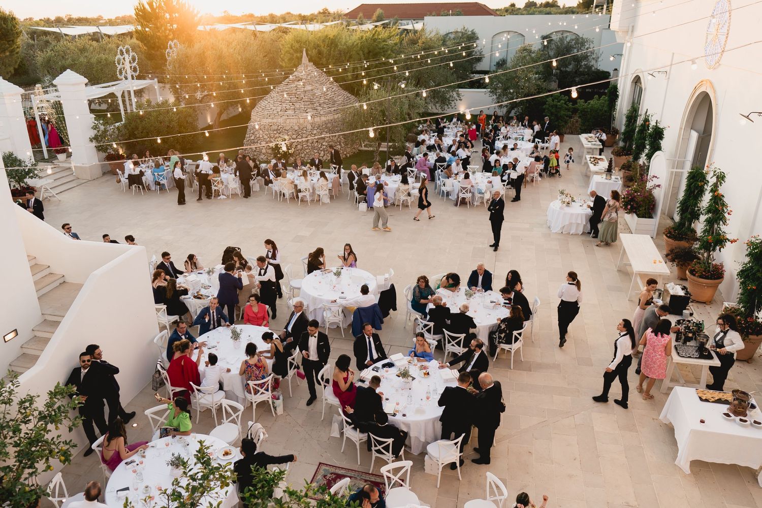 Aerial view of outdoor wedding reception with guests dining under string lights at dusk.
