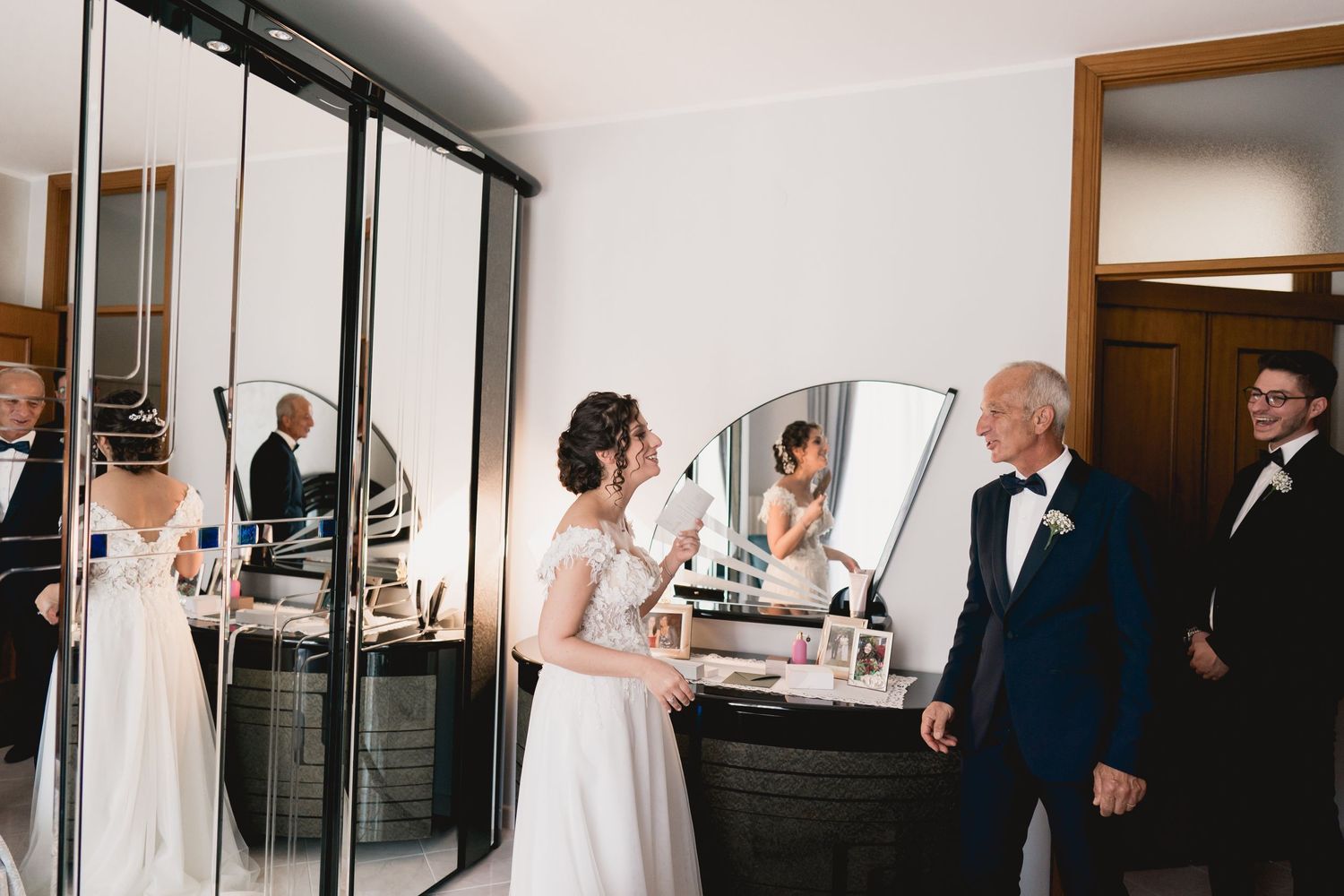 Wedding party members gather around a vanity mirror in a getting-ready room.