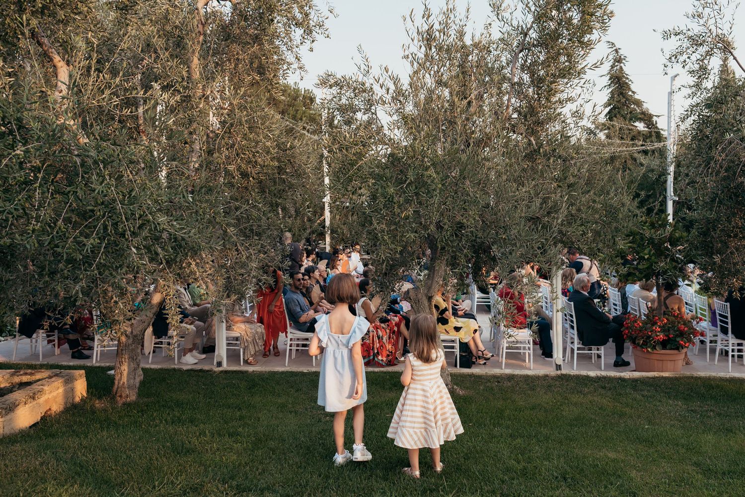 Two small children in white dresses dance on grass during an outdoor wedding celebration.