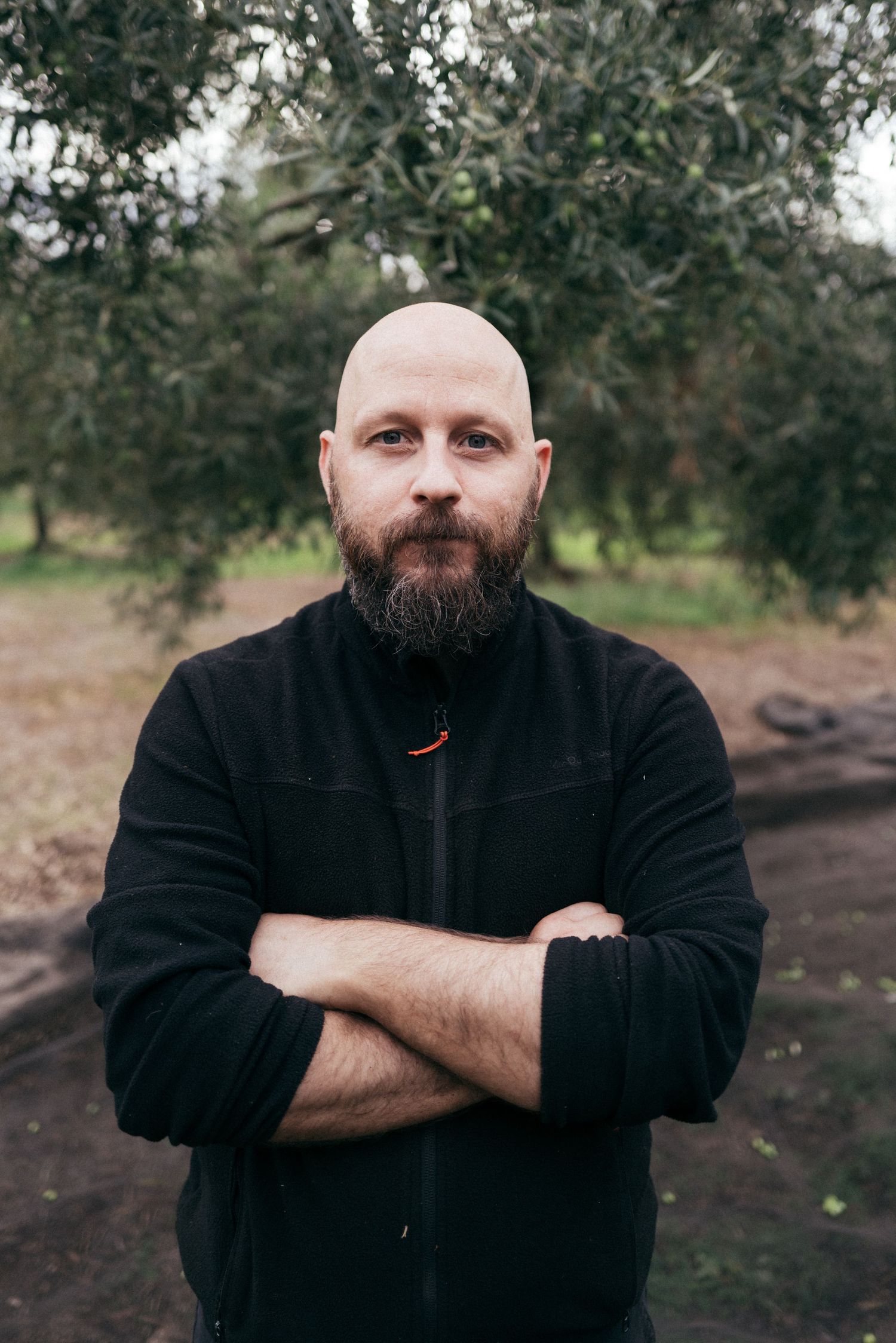 Person in black jacket standing in olive grove with crossed arms.