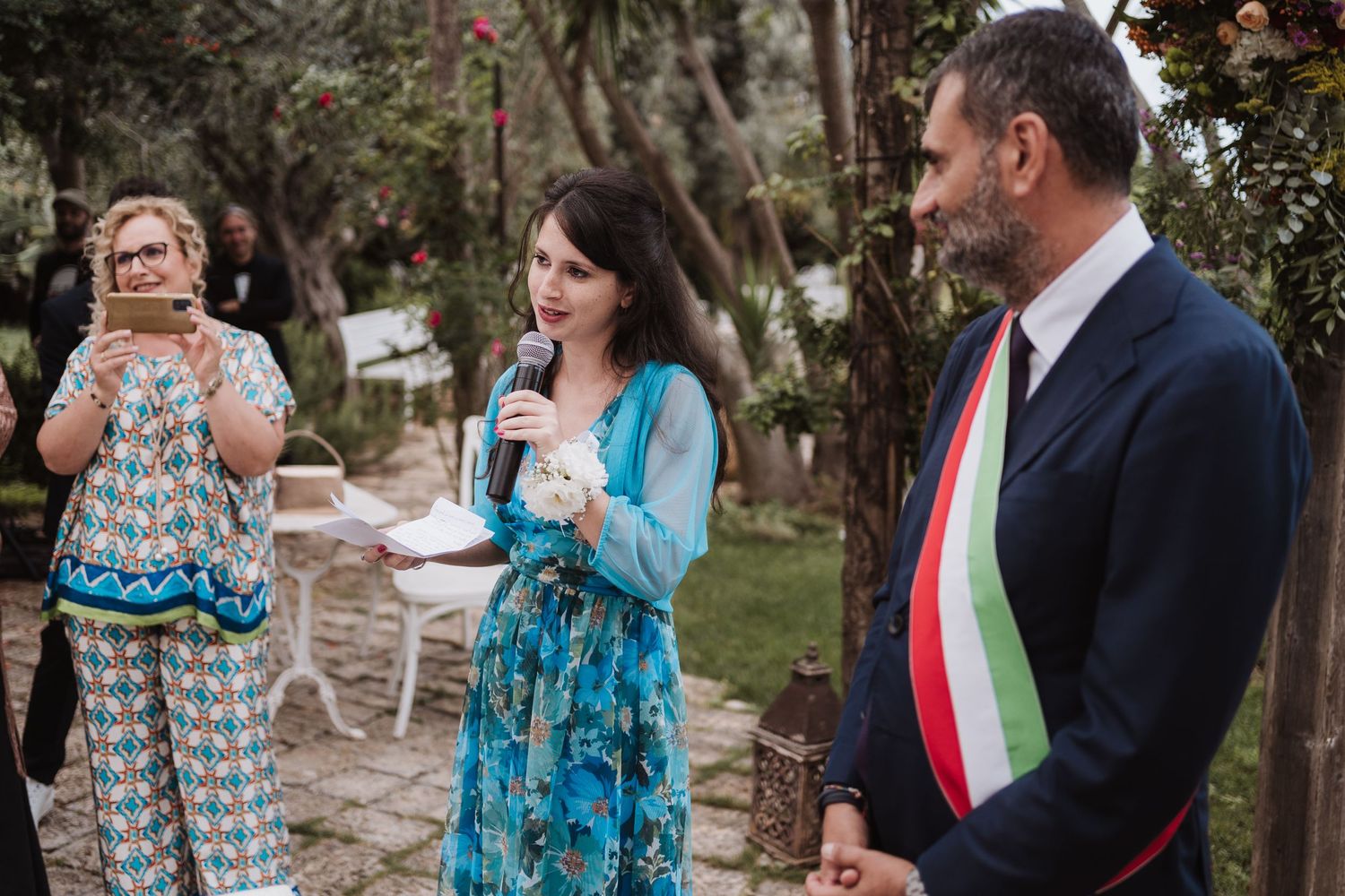 Guests and official in ceremonial sash participate in outdoor wedding proceedings.