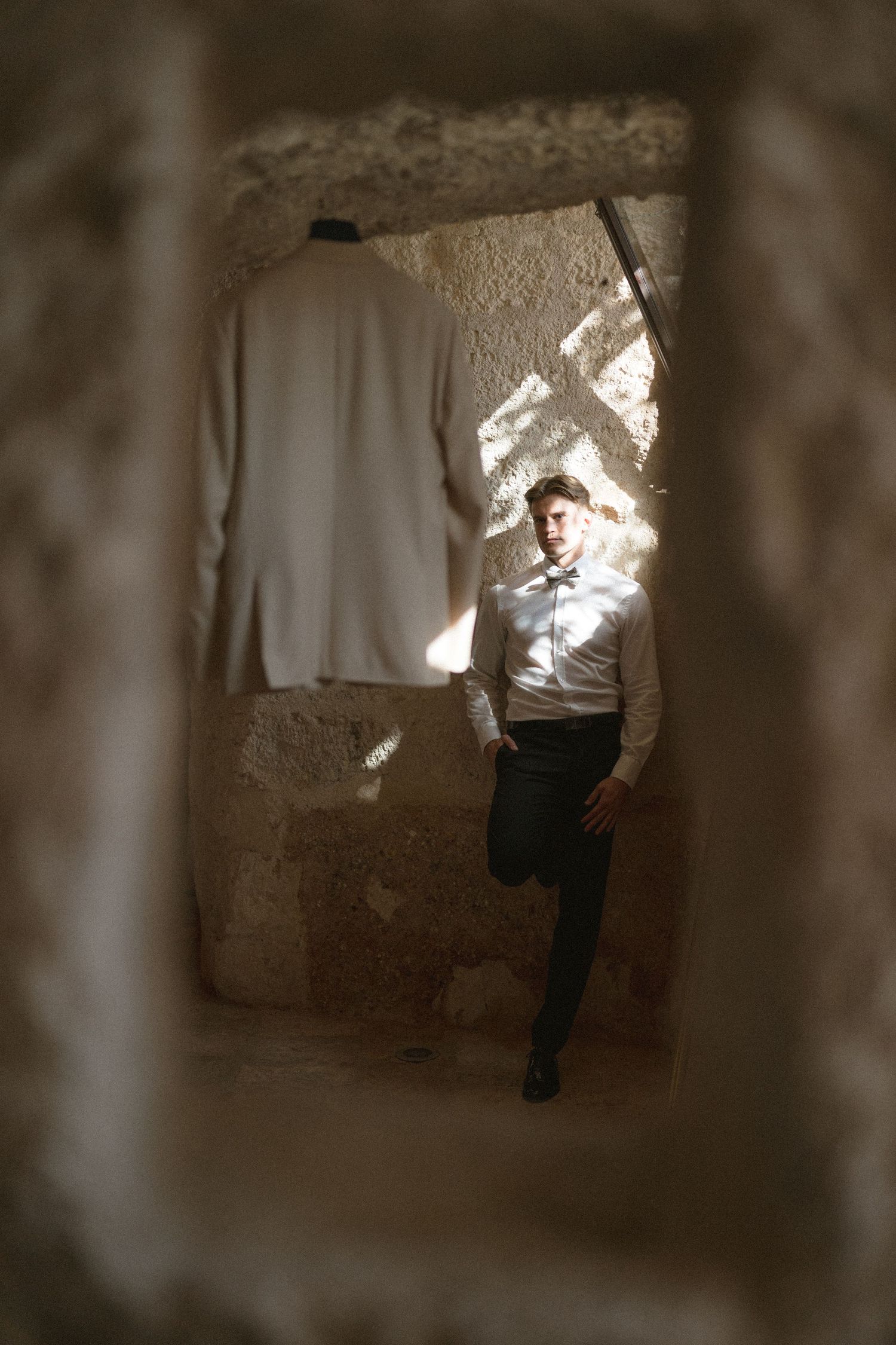 Groom in a white shirt and bow tie leans against a sunlit stone wall, framed by a rough rock opening, with a white jacket hanging nearby.