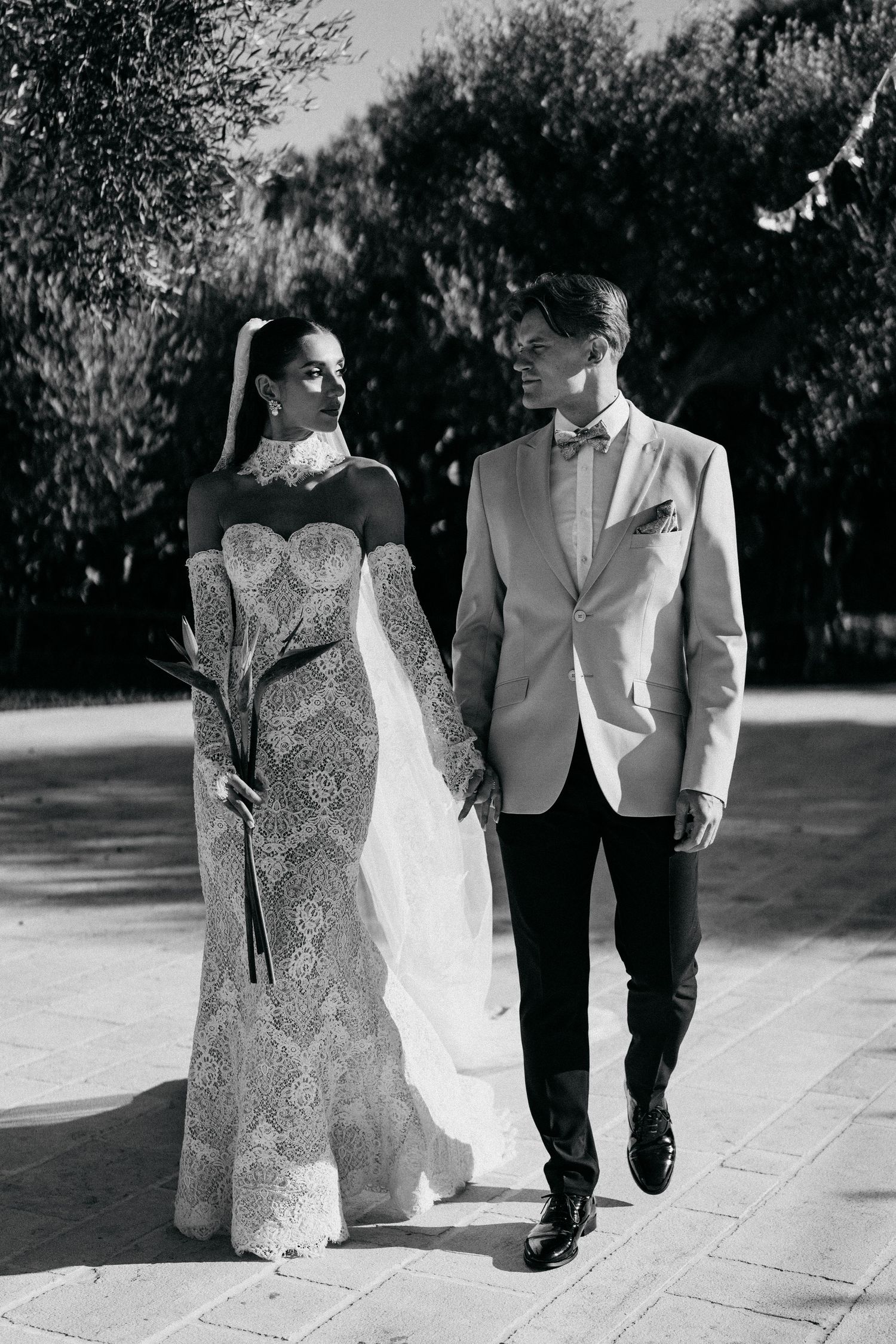 Black and white photo of a bride in a lace gown and a groom in a light blazer walking hand in hand along a tree-lined path.